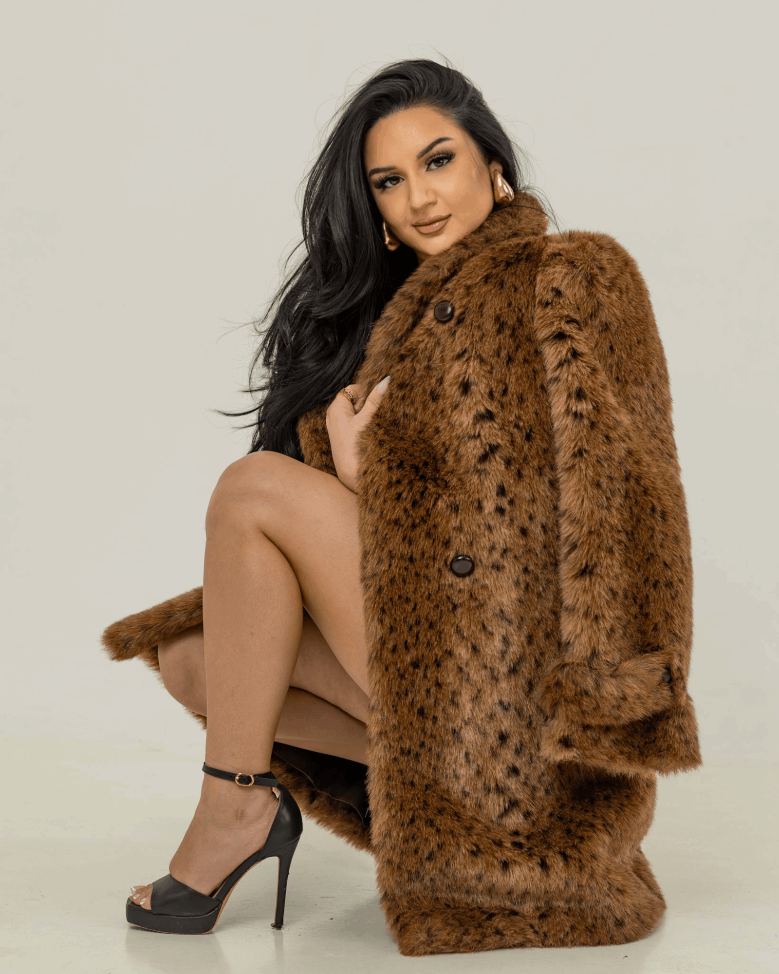Woman with long dark hair wearing a brown fur coat, squatting with one knee up, against a plain background.