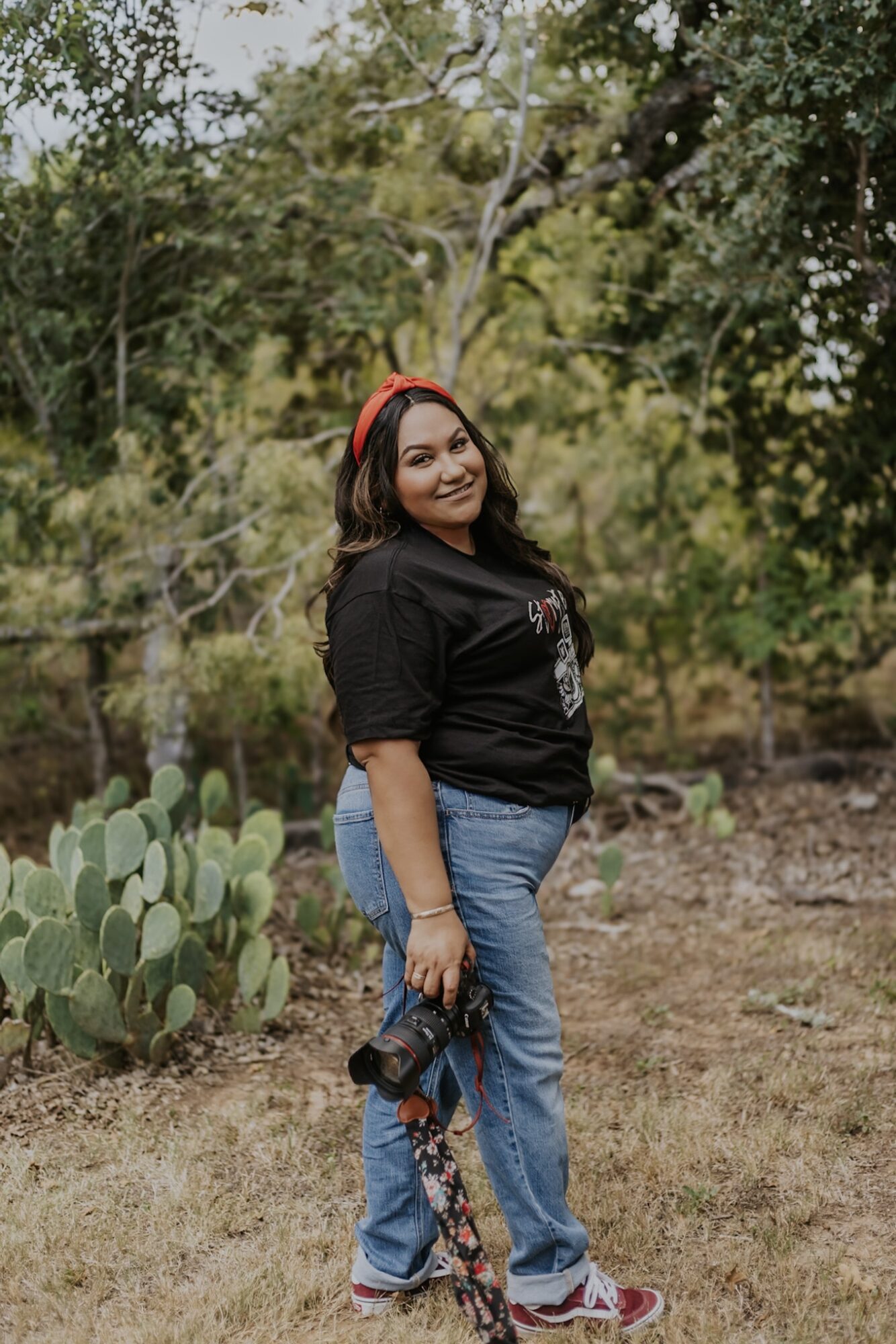 Young woman with red headphones standing outdoors near cactus plants, smiling, with trees in background.
