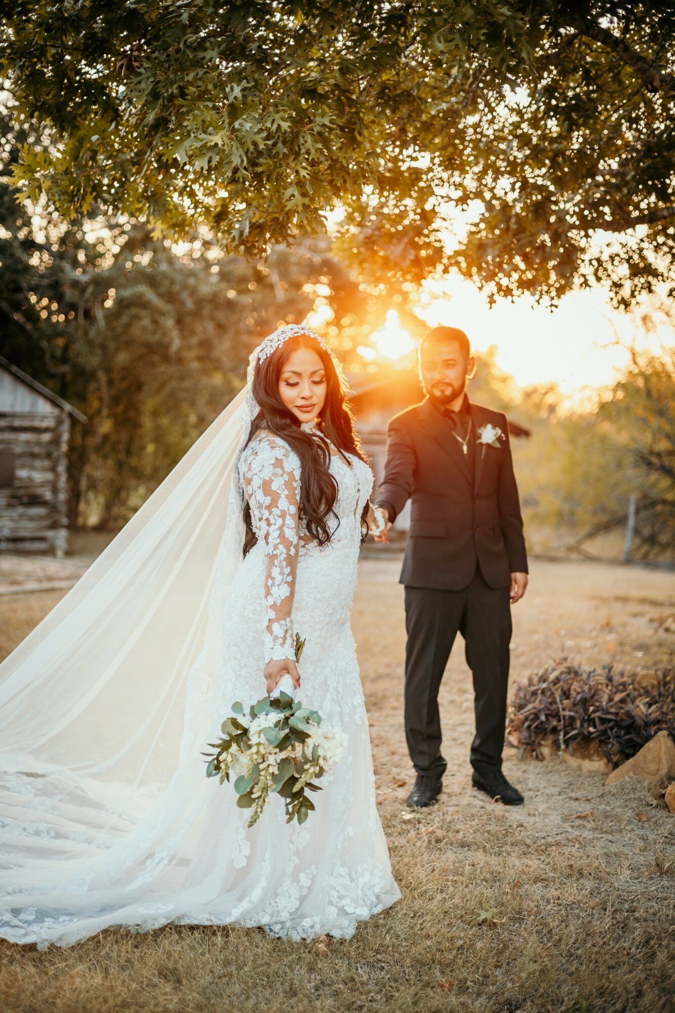 Bride in a white lace wedding dress and veil holding a bouquet, standing outdoors with groom in a suit, sunset background.