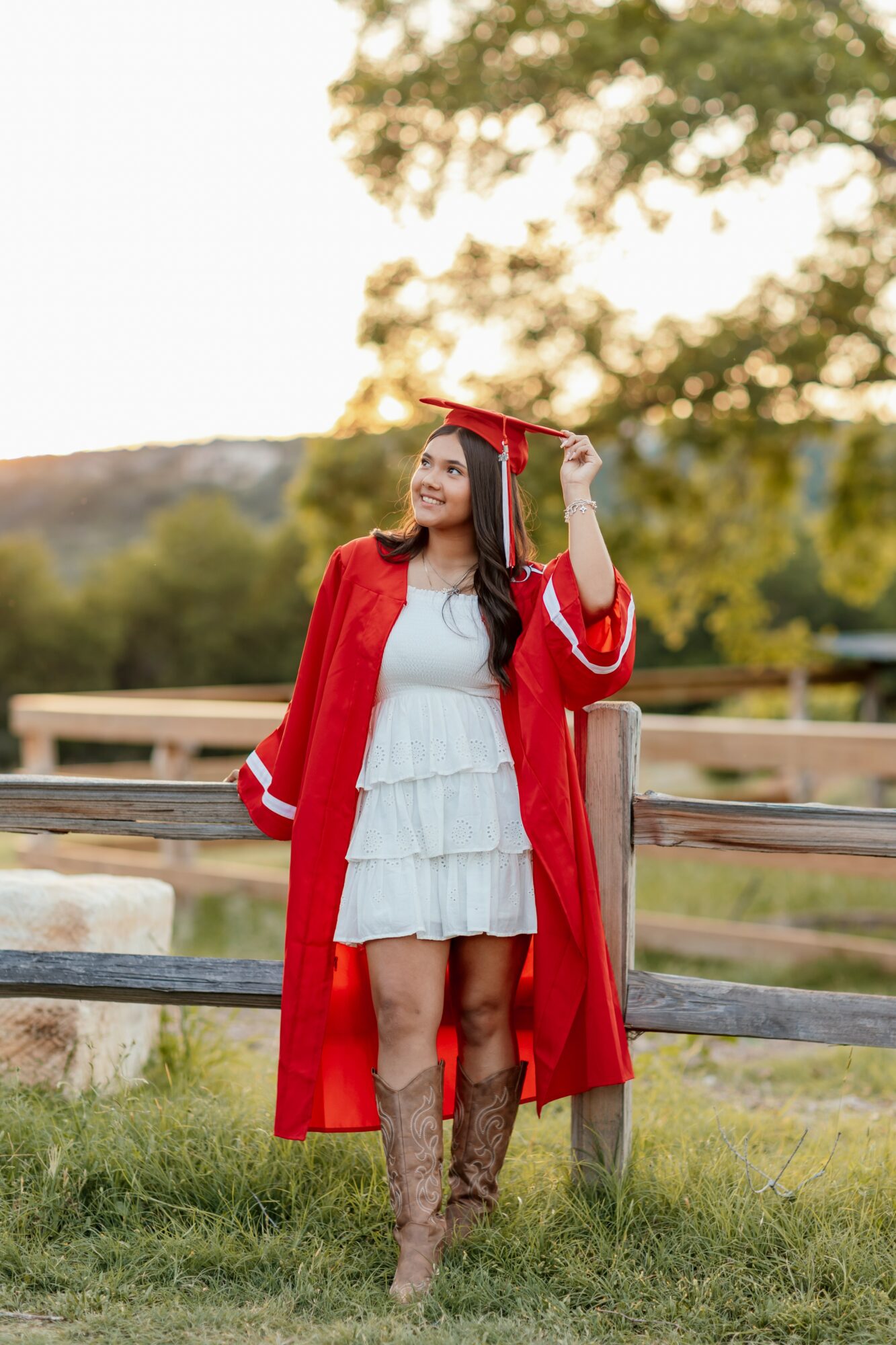 Young woman in graduation gown and cap standing outdoors near a wooden fence, smiling, with trees and sky in background.