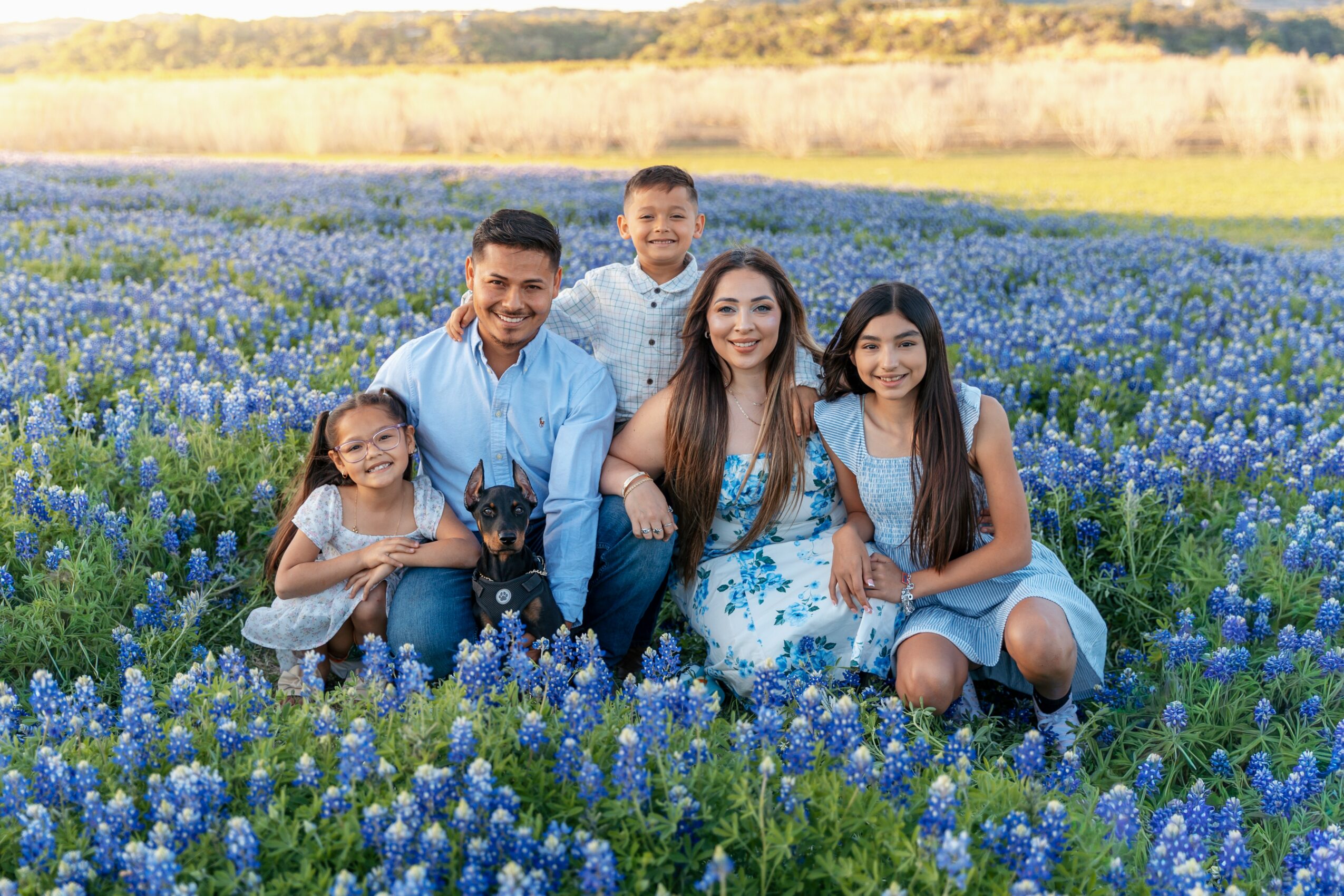Family of five sitting in a field of blue flowers, smiling, with a landscape background.