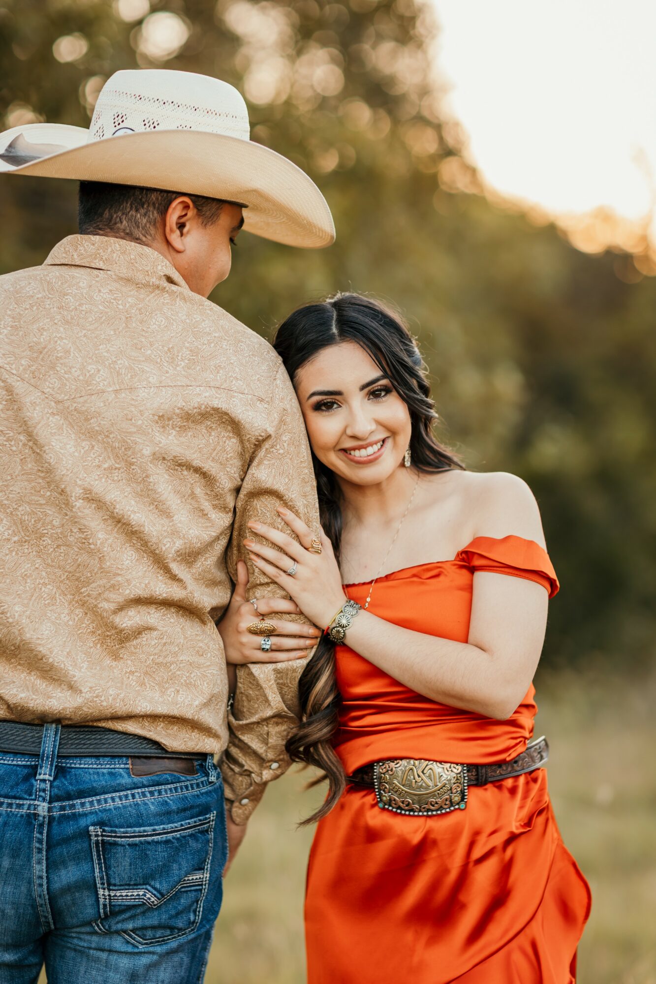 A woman in an orange dress smiling and holding a man's arm outdoors during sunset.