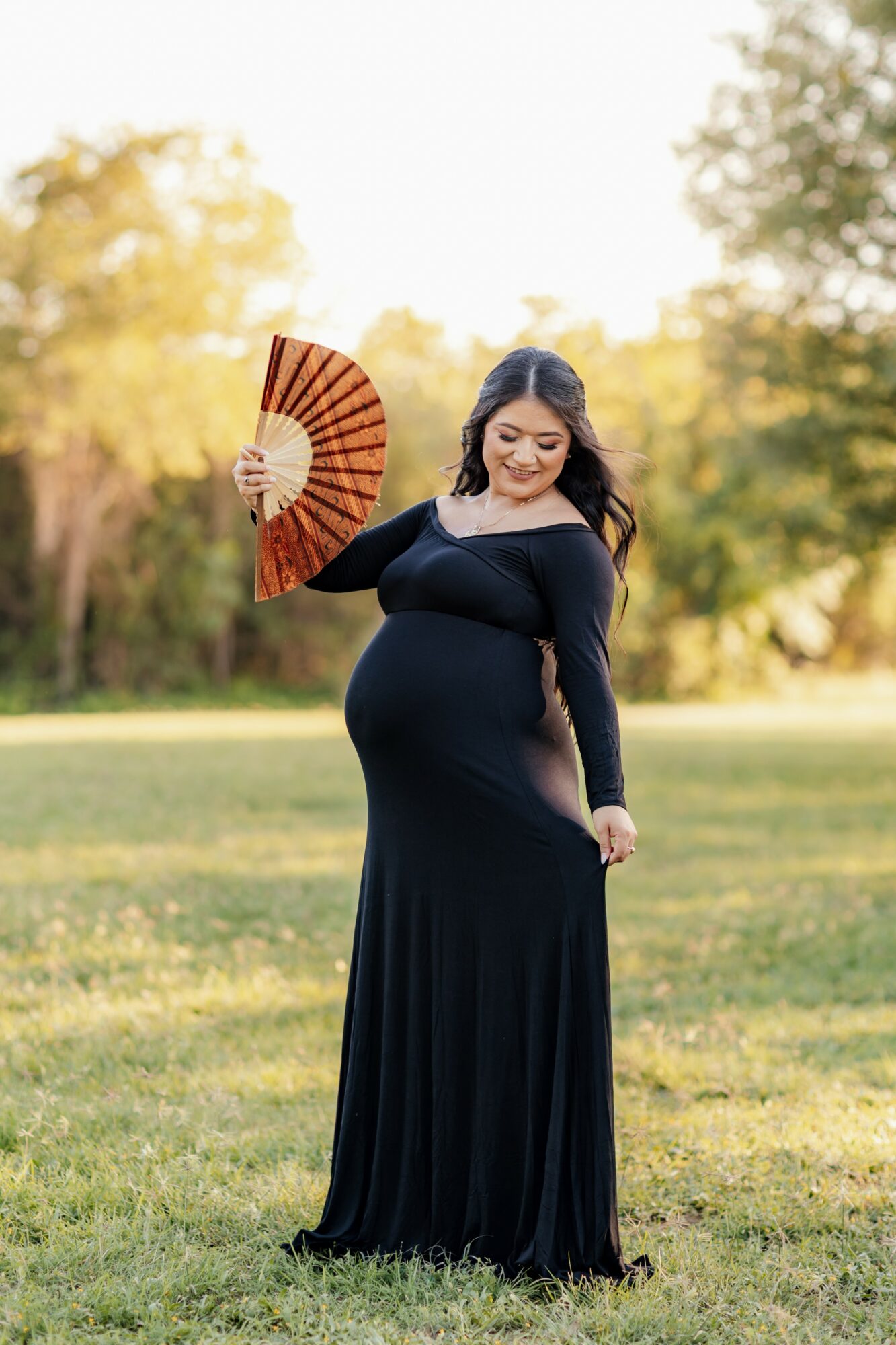 Pregnant woman in a long black dress holding a fan outdoors, smiling, with trees and grass in the background.