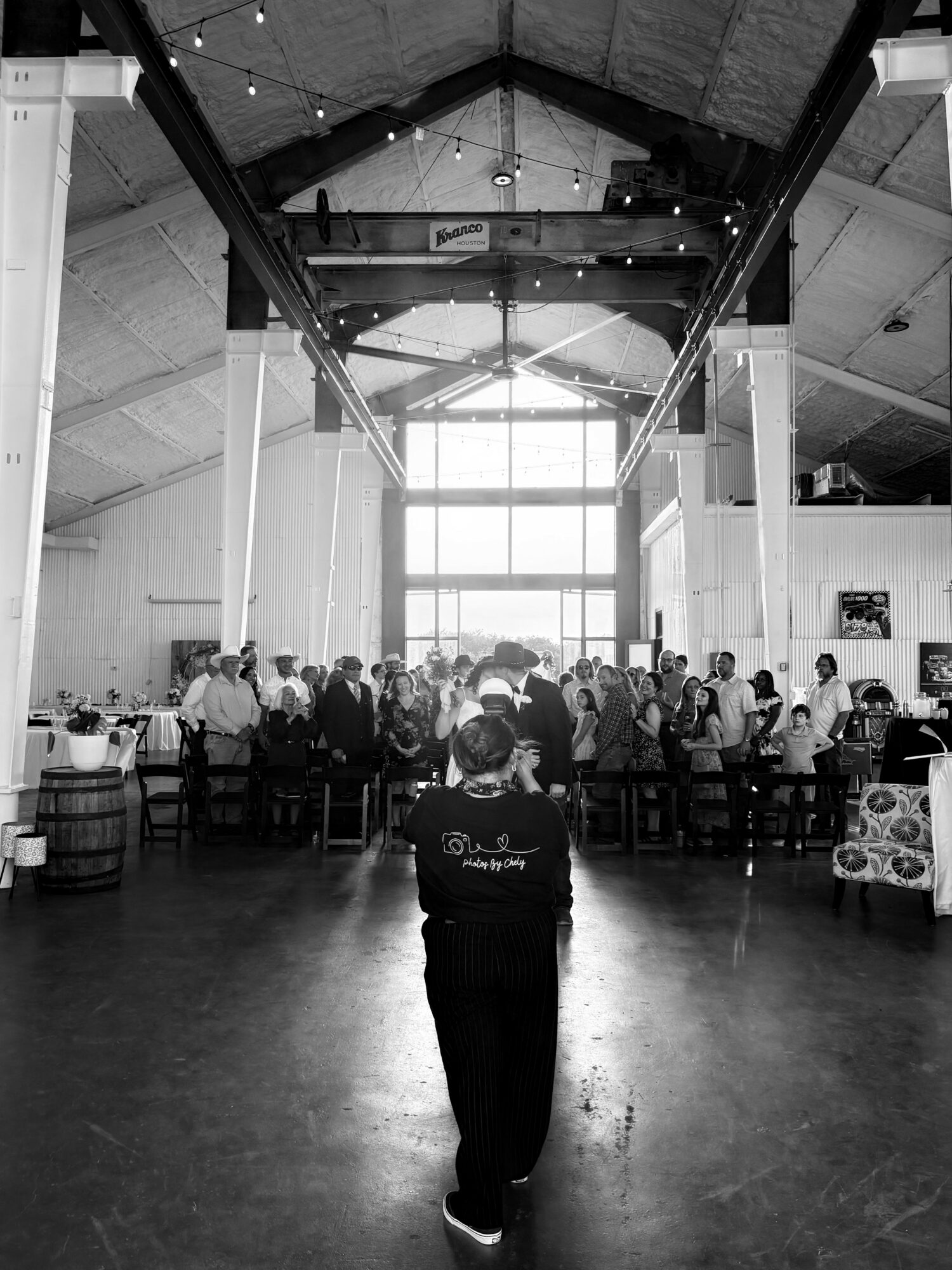 Person standing in a large indoor space with high ceiling and large windows, facing a group of people gathered around tables.