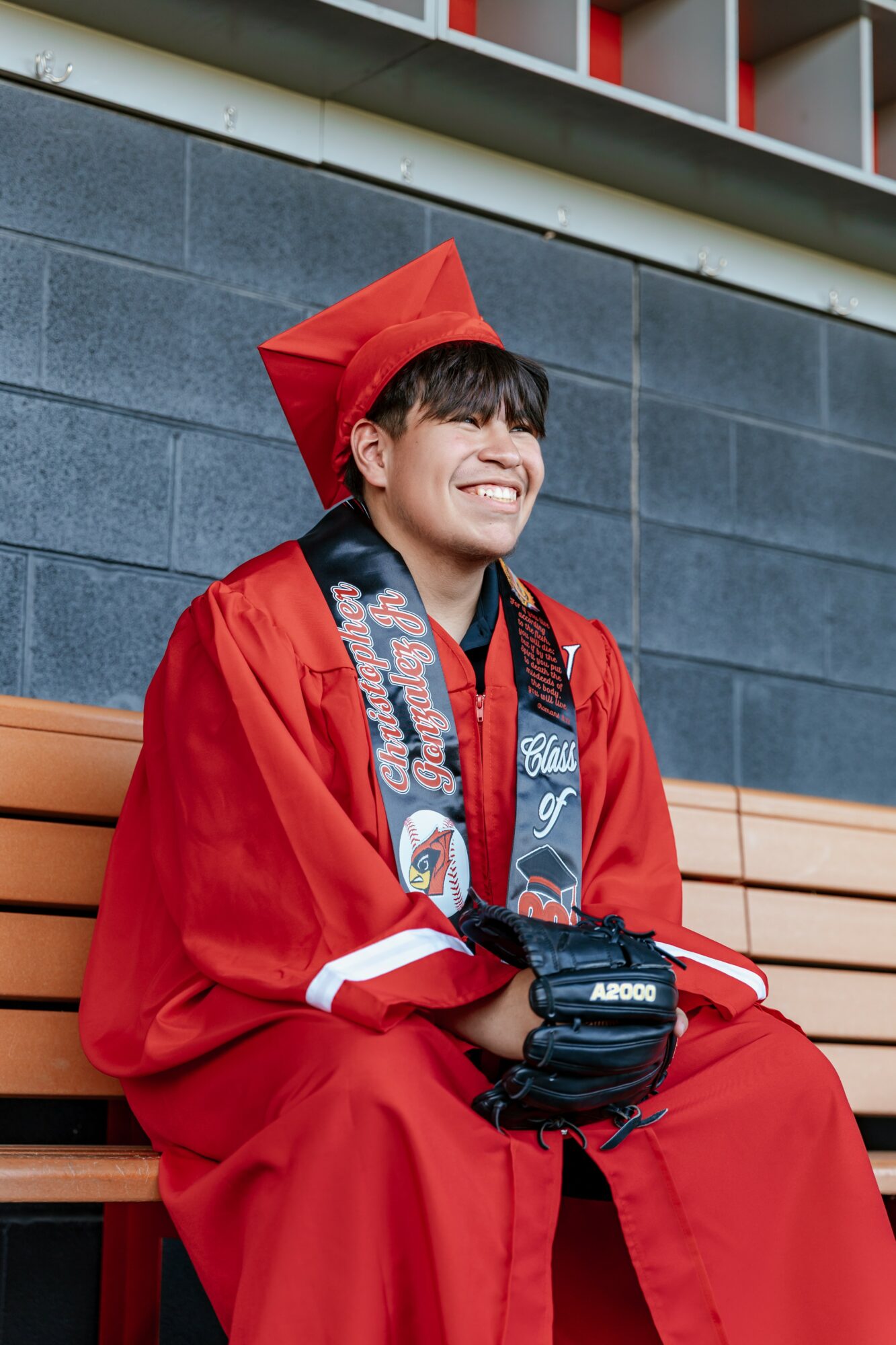 Young person in red graduation gown and cap sitting on a bench, smiling, holding a diploma, against a gray wall.
