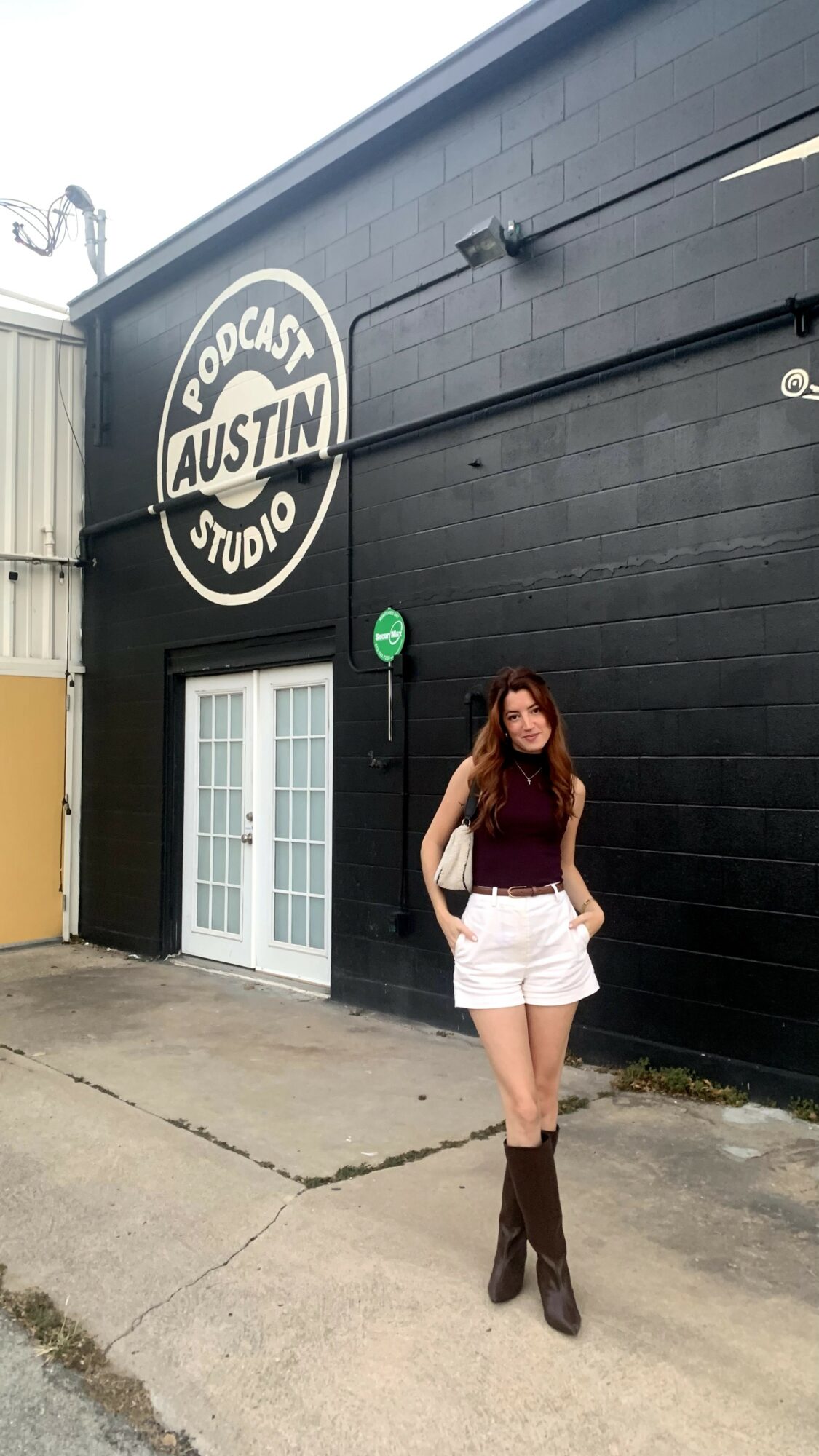 Woman with long hair wearing a sleeveless top, white shorts, and knee-high boots standing outside a building with a sign that reads 'Podcast Austin Studio'.