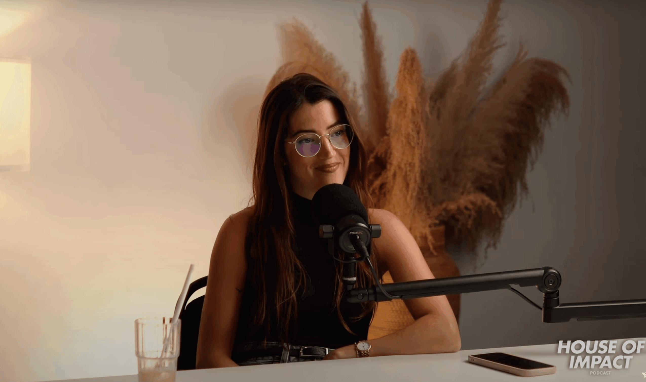 Woman with long hair and glasses speaking into a microphone at a desk, with pampas grass in the background.