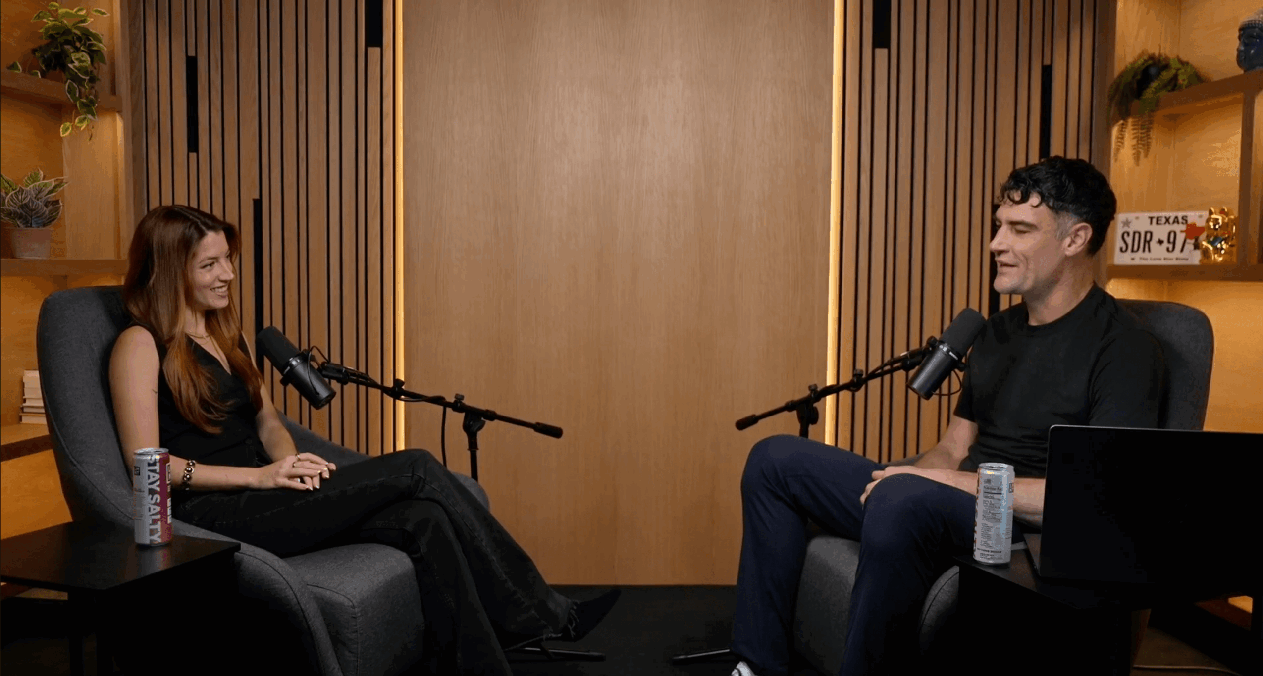 Two people sit across from each other in a recording studio, speaking into microphones, with drinks on small tables nearby.