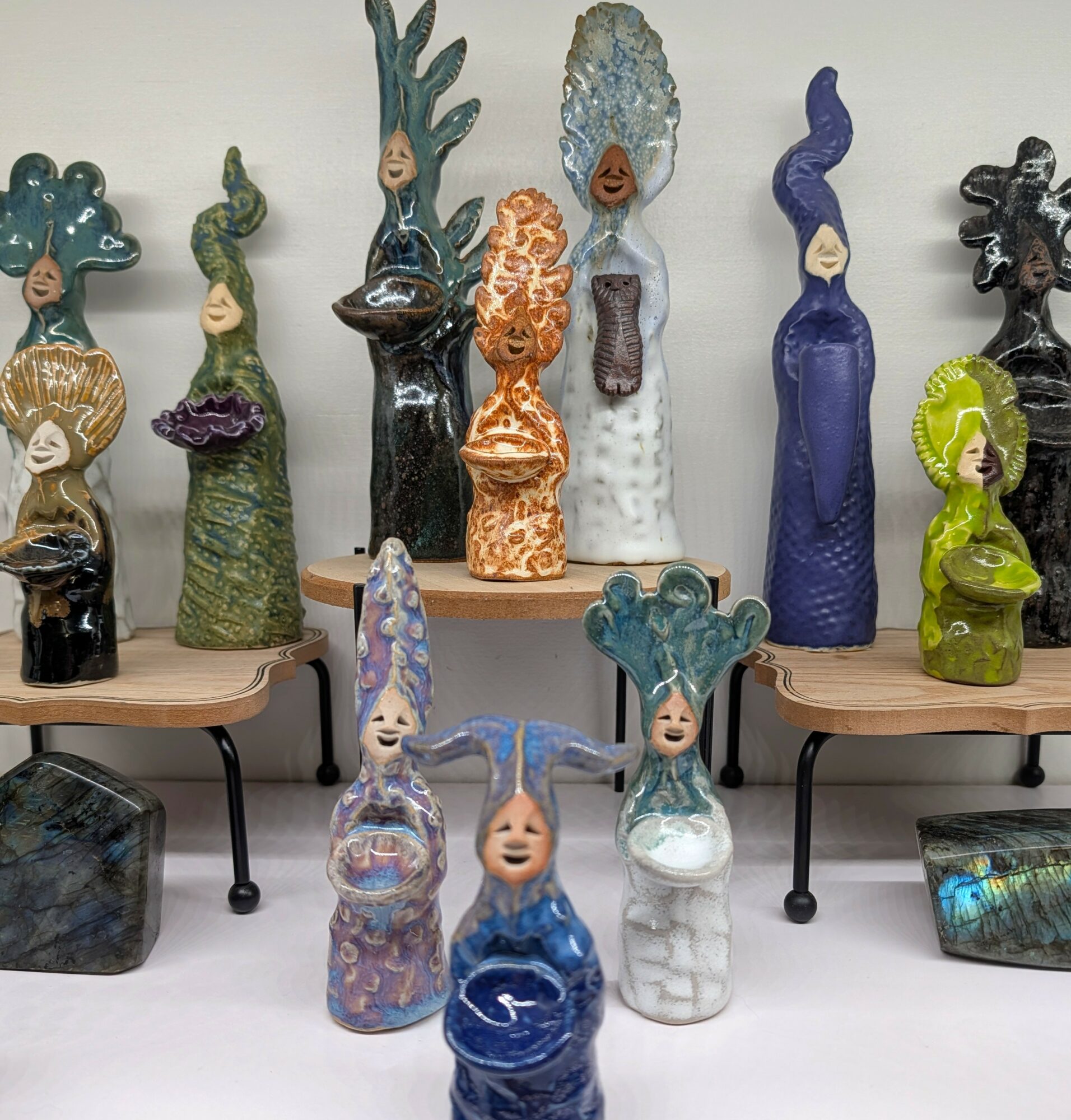 Collection of colorful ceramic sculptures with faces and elaborate headpieces, displayed on tables and stands, against a plain background.