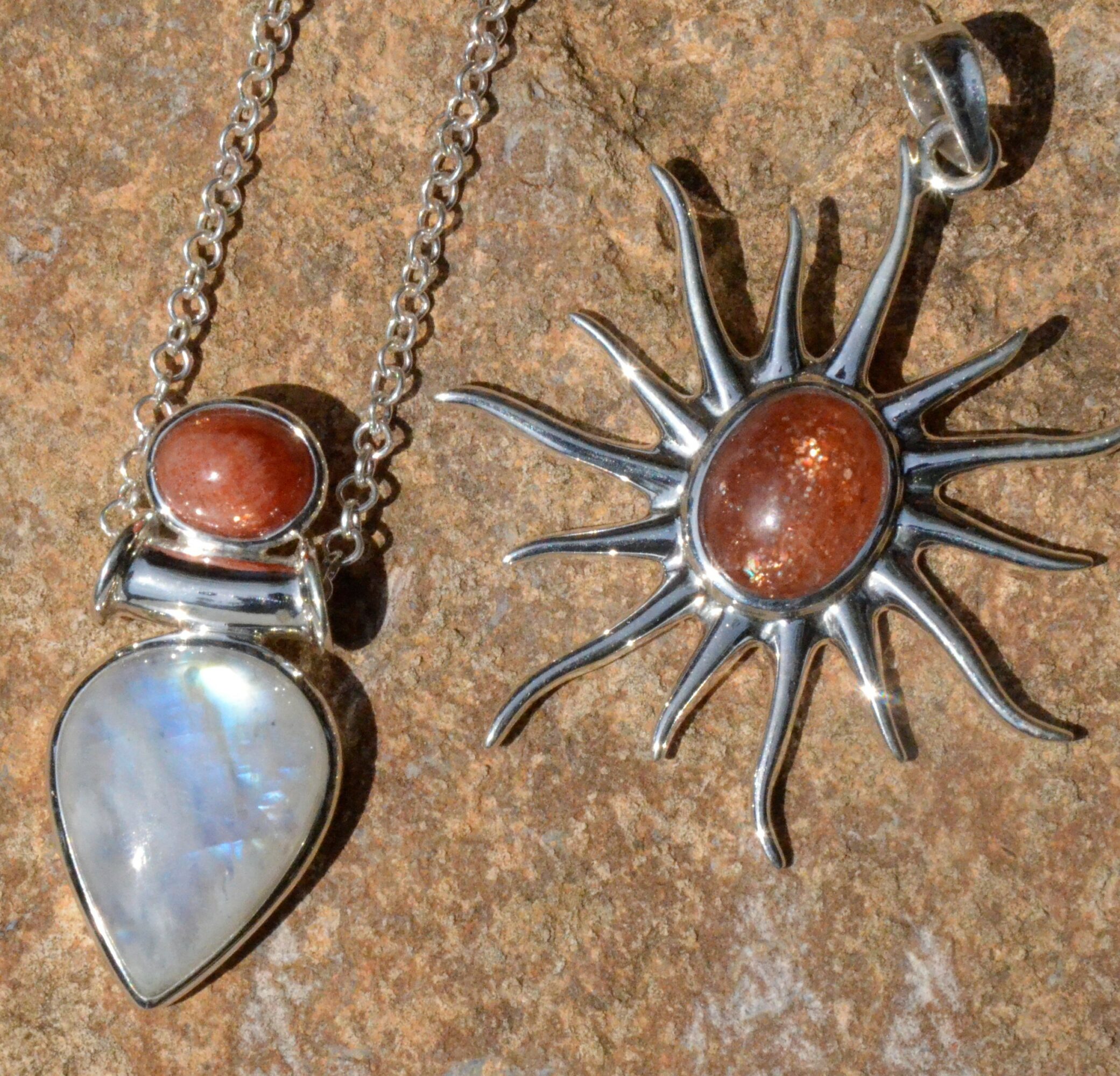 Two silver necklaces with sun and heart-shaped pendants on a textured surface.