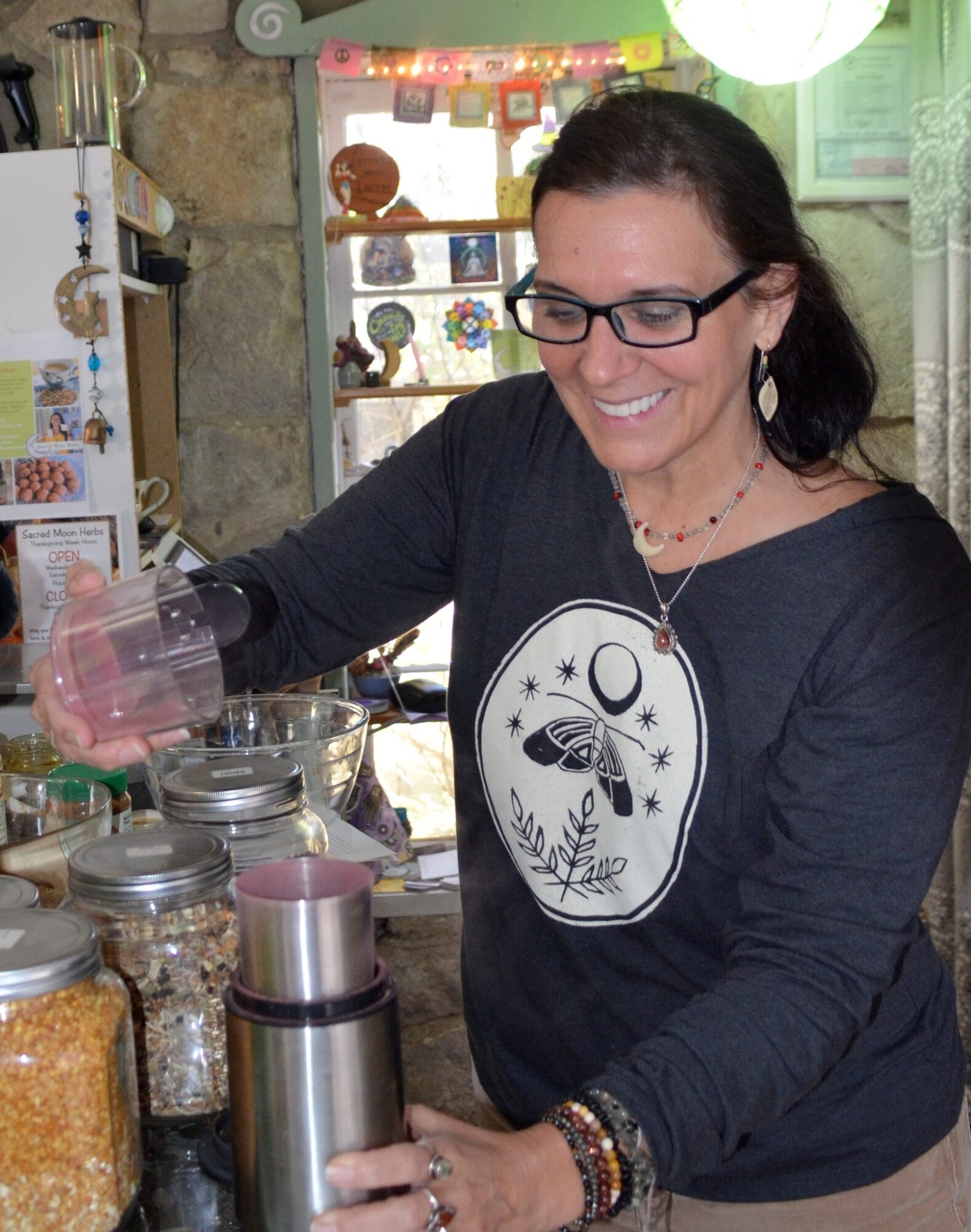 Woman with glasses smiling, preparing food in a market stall with jars and containers, wearing a black sweatshirt with a moon and butterfly design.