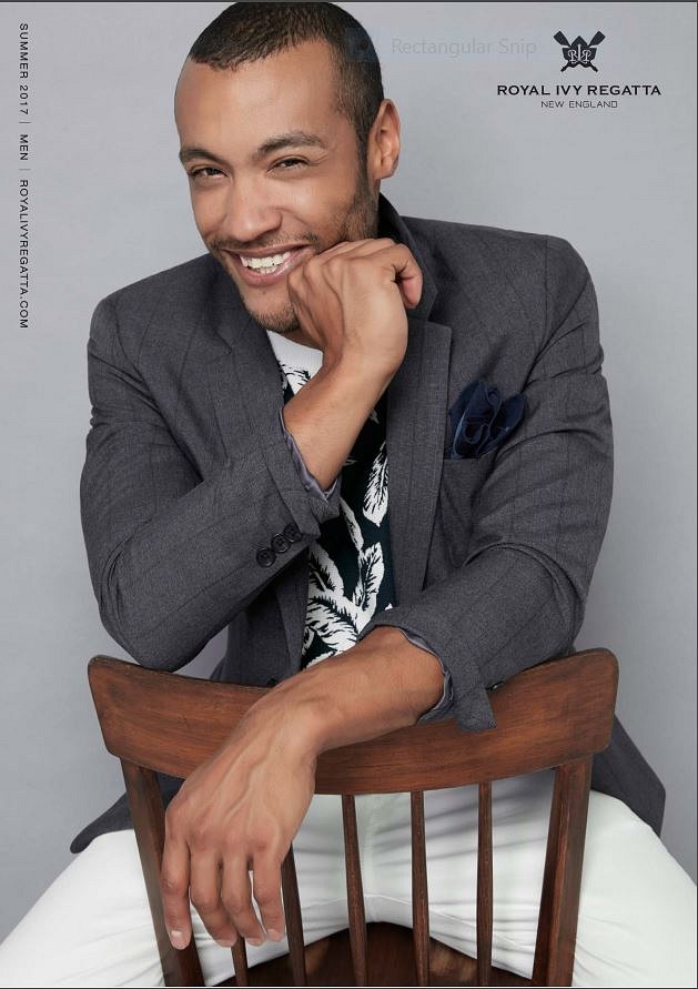 Smiling man in a gray blazer sitting on a wooden chair, with a light gray background and event logo in top right corner.
