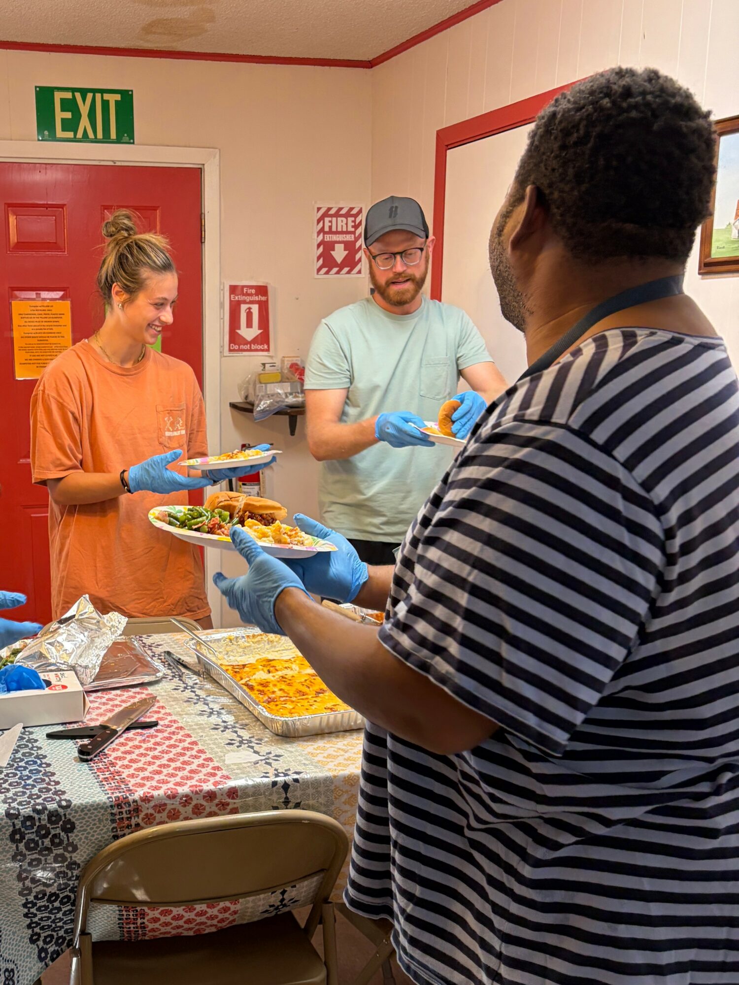 Three people serving food at a table, one woman and two men, in a room with a red door and exit sign.