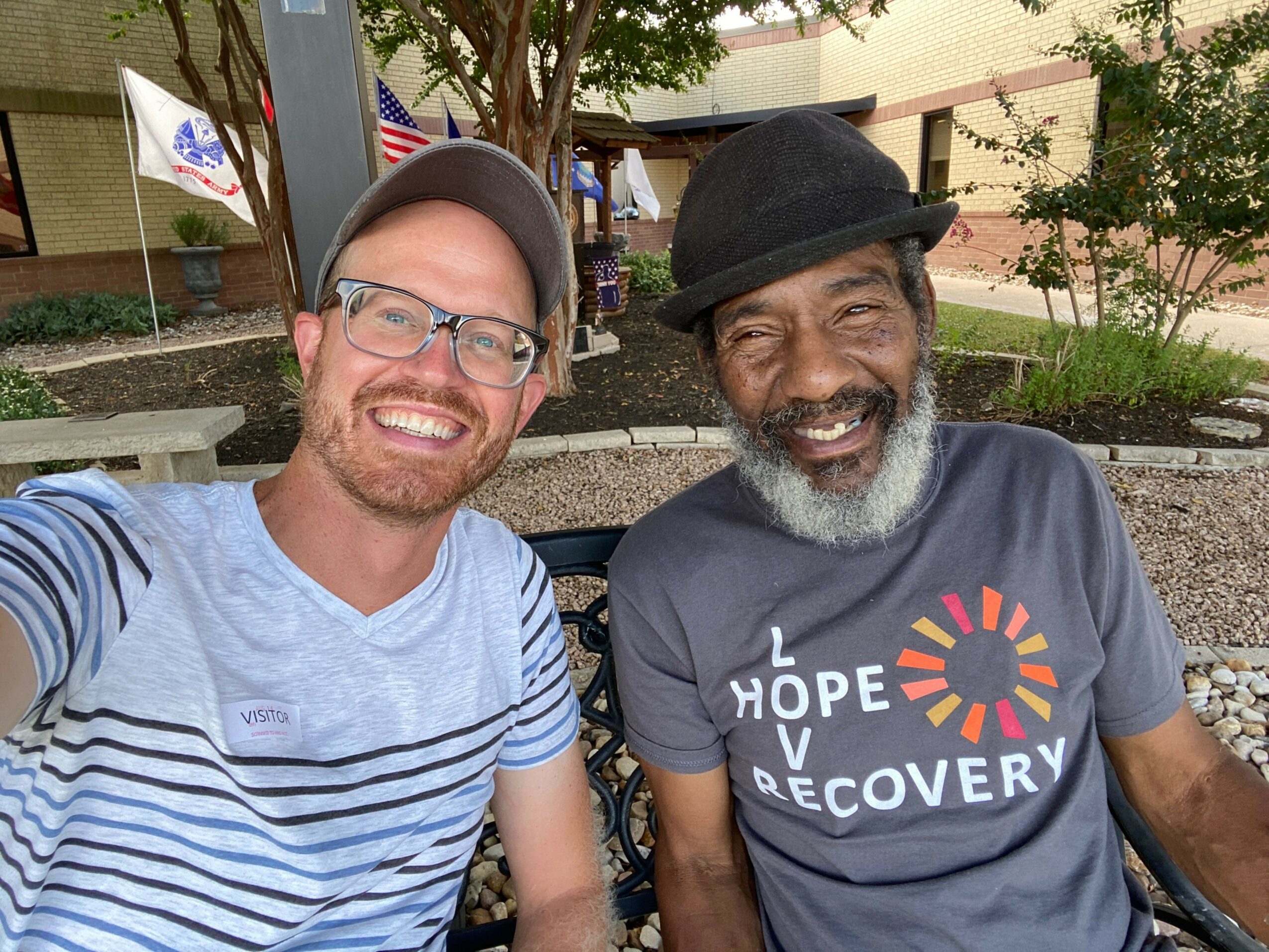 Two men smiling, one with a beard and glasses, the other with a hat, standing outdoors near a building.