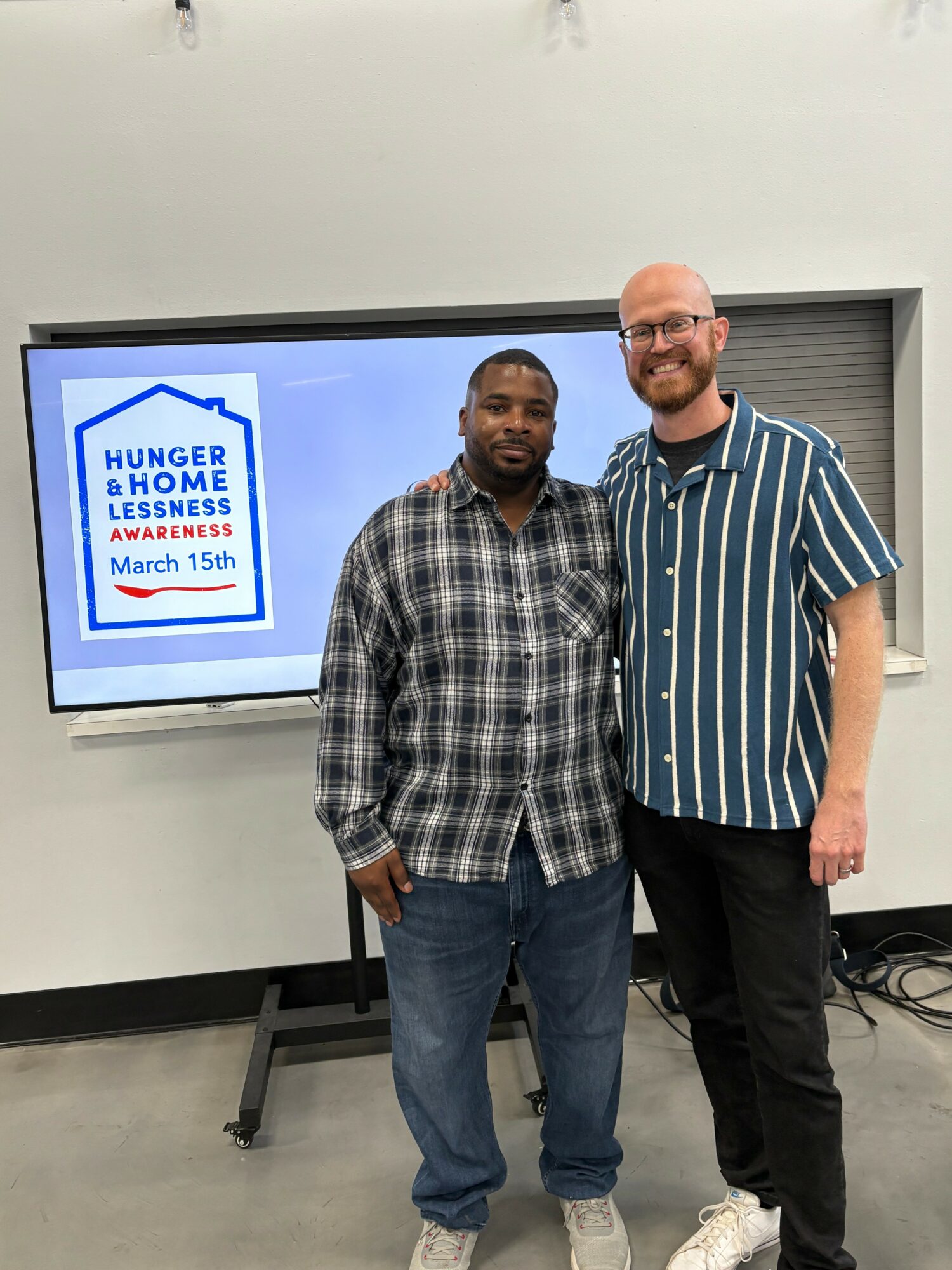 Two men standing side by side in front of a screen displaying a logo and text about hunger and homelessness awareness, smiling.