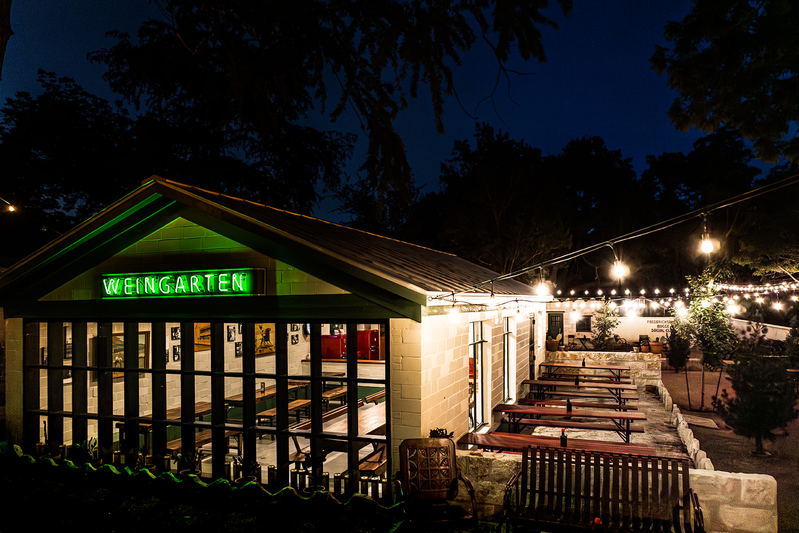 Night view of a building with a green illuminated sign reading 'WEINGARTEN', outdoor seating area, string lights, and trees in background.