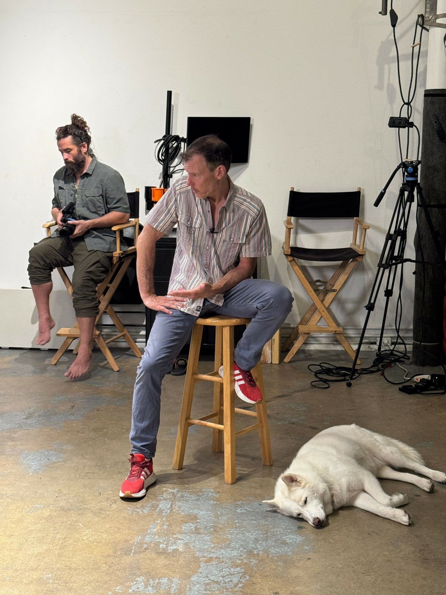 Two people sitting on chairs and a dog lying on the floor indoors, with equipment in the background.