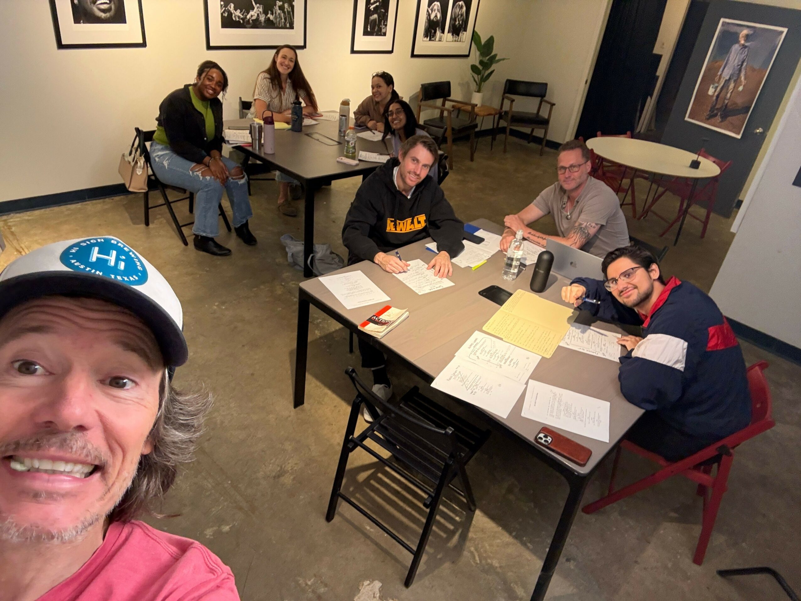 Four people sitting at a table with papers and drinks, two standing, one lying on the floor, one taking a selfie.