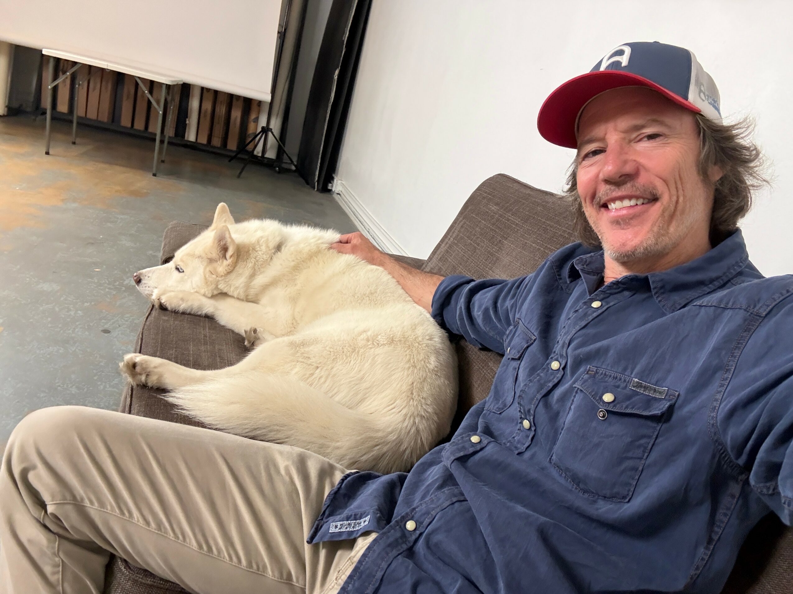 Man sitting on a couch with a white dog lying beside him, smiling, indoors.