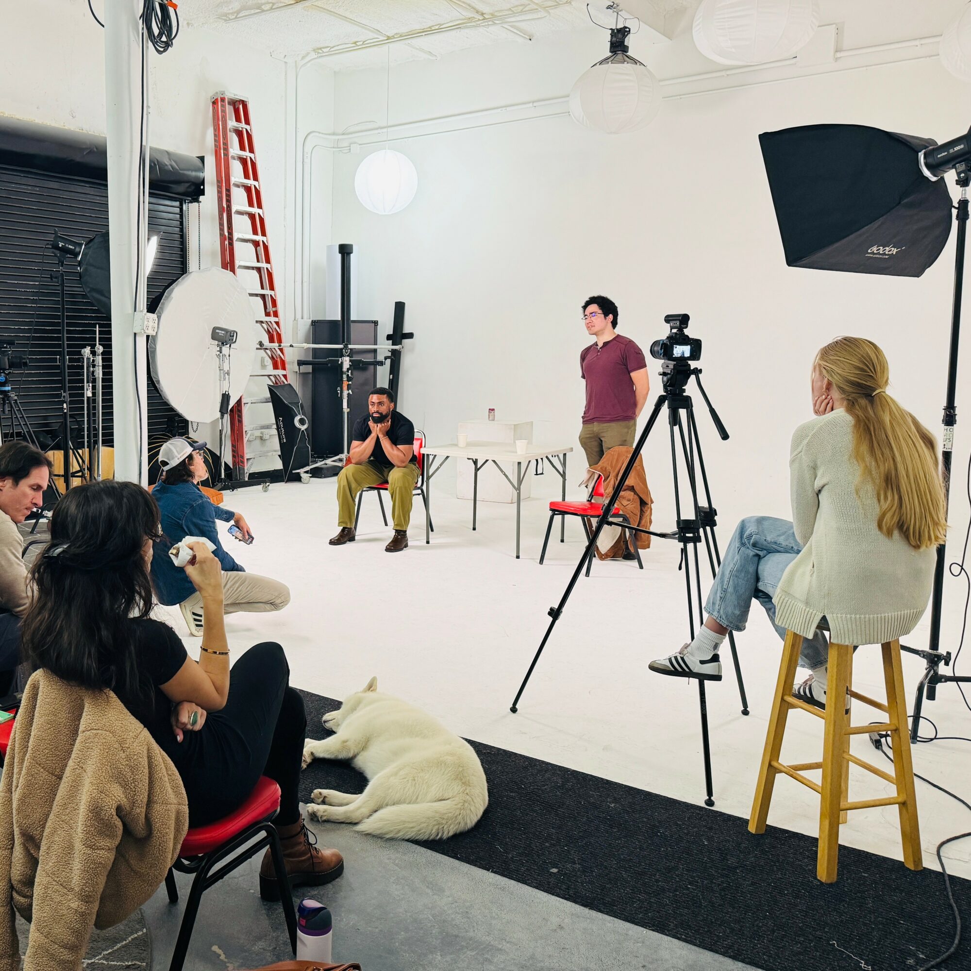 People in a photography studio with equipment, some seated and some standing, during a photo shoot or interview, with a white background.