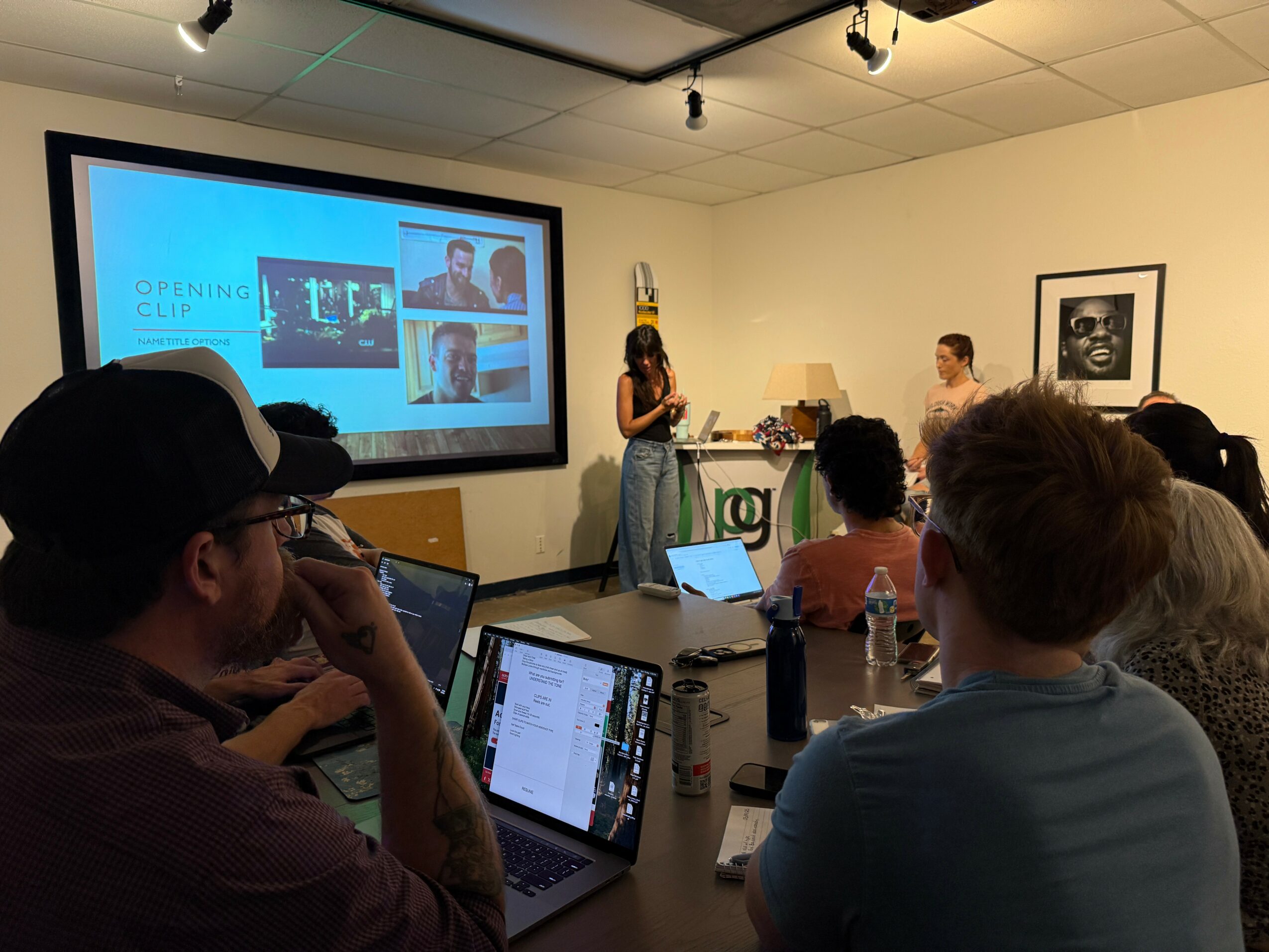 A woman presents in front of a screen with images, audience seated at tables, some using laptops, in a room with framed art.