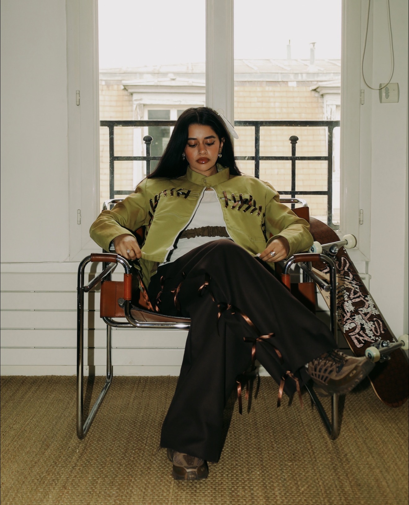 Woman with dark hair and makeup sitting on a bench in front of a window, wearing a green jacket and black pants.