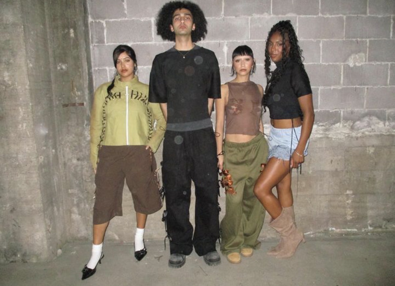 Four people standing against a concrete wall, with varied clothing styles and poses.