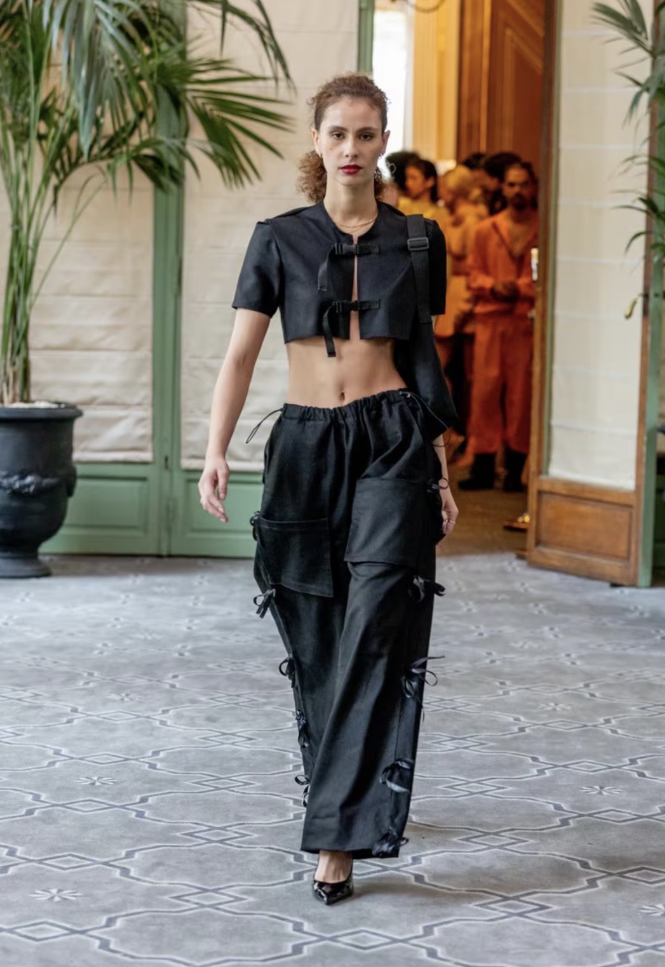 Woman walking outdoors in black crop top and baggy pants, with people in background, plants, and open door behind her.
