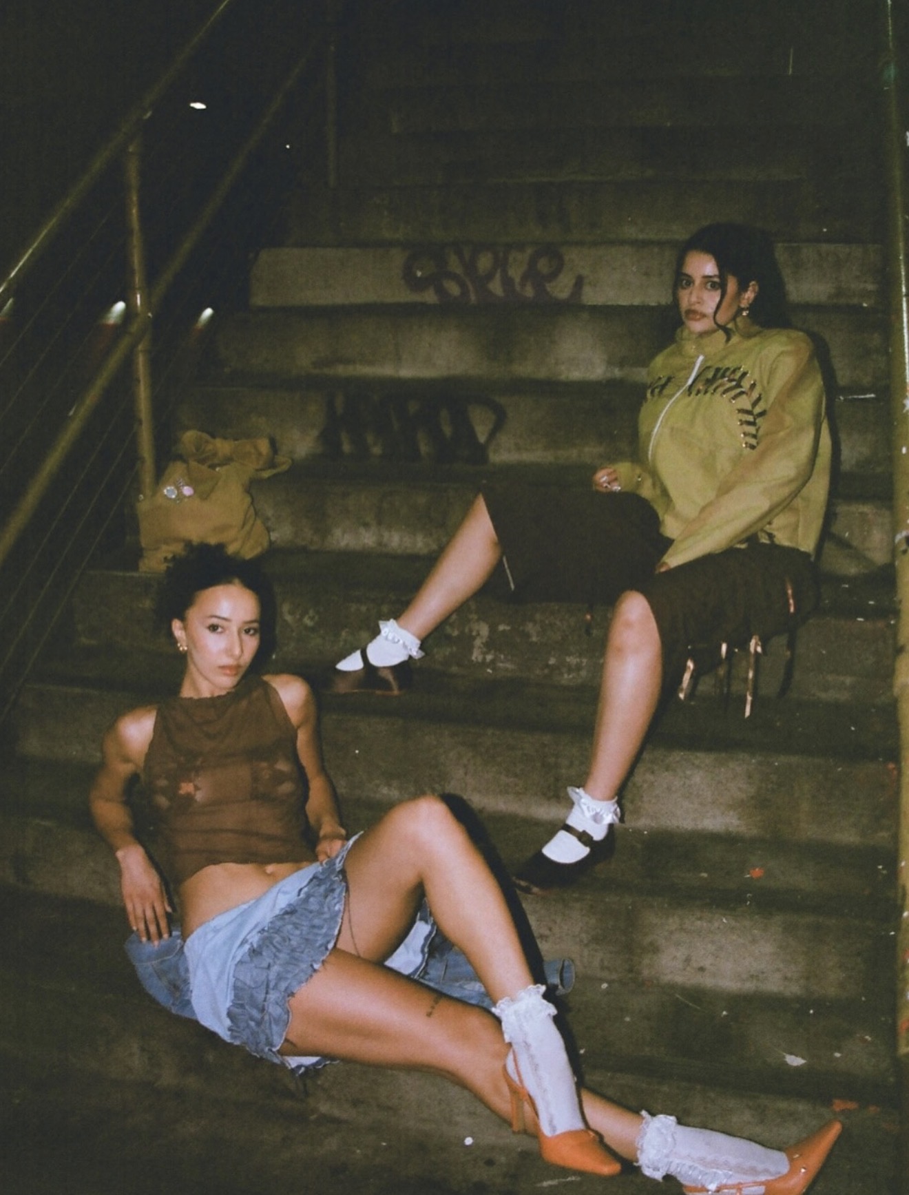 Two women sit on stairs in a dimly lit setting, one leaning against the wall, the other reclining on the steps.