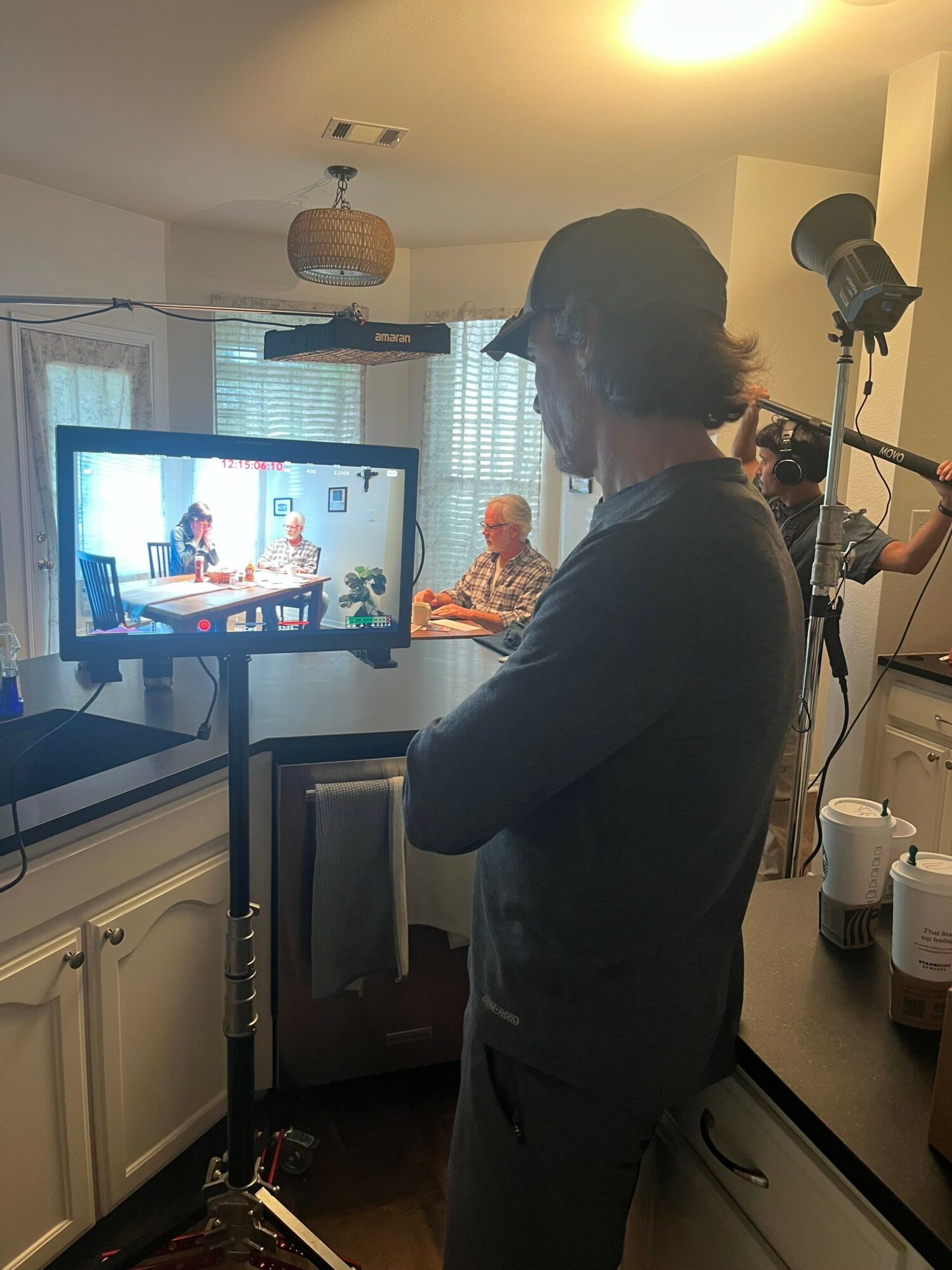 Person with dark hair in a ponytail, wearing a dark shirt, standing in a kitchen, looking at a computer monitor.