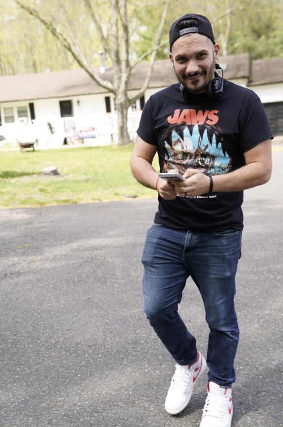 Man standing outdoors on a paved surface, holding a phone, wearing a black Jaws t-shirt, jeans, and white sneakers.
