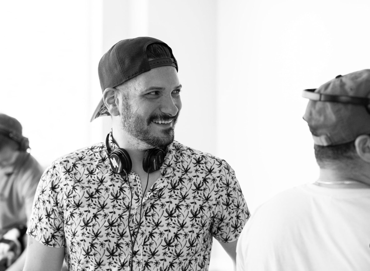 Smiling man with a beard, wearing a patterned shirt and headphones around his neck, talking to another person.