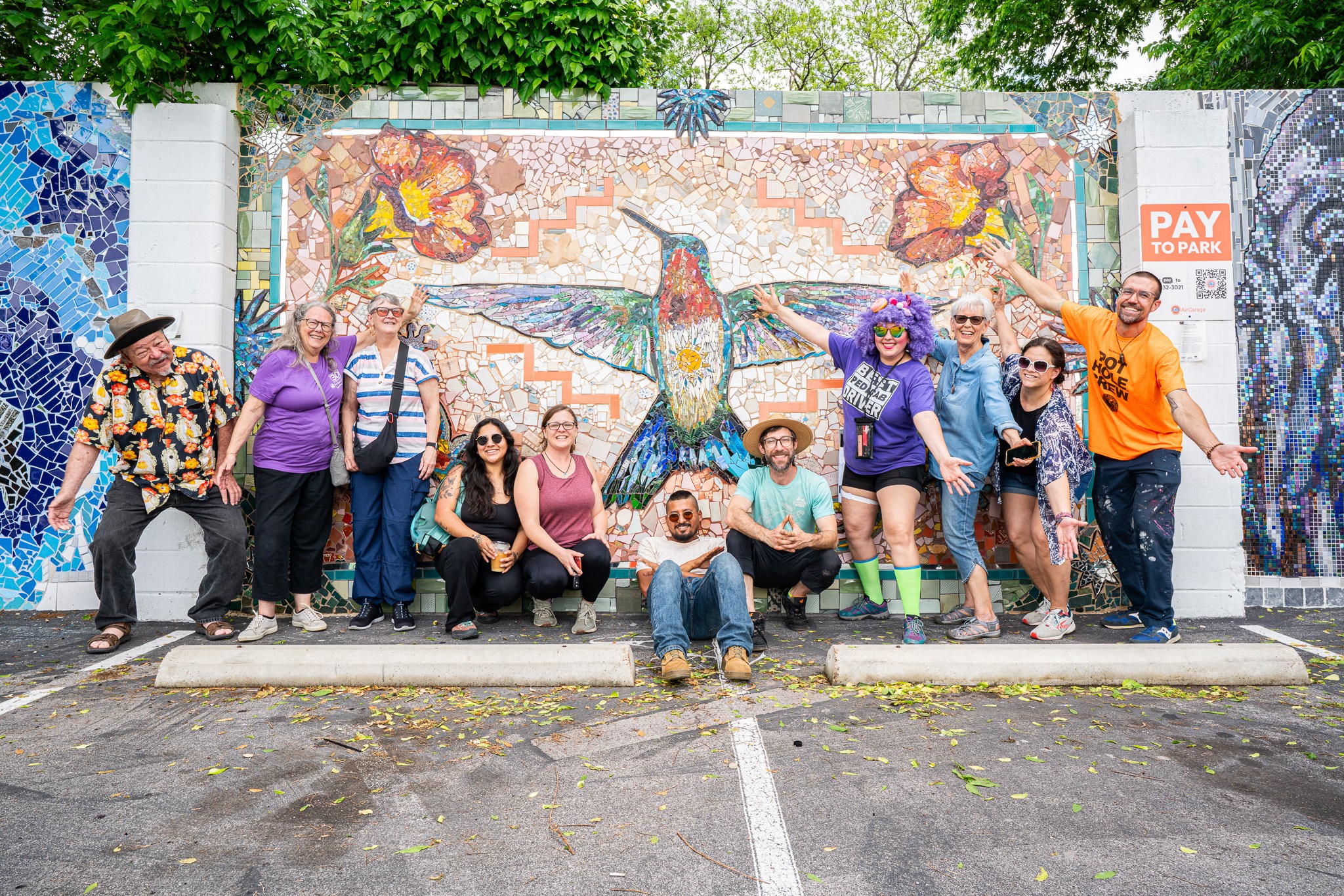 Group of people posing in front of a colorful mosaic mural, some sitting and some standing, outdoors on pavement.