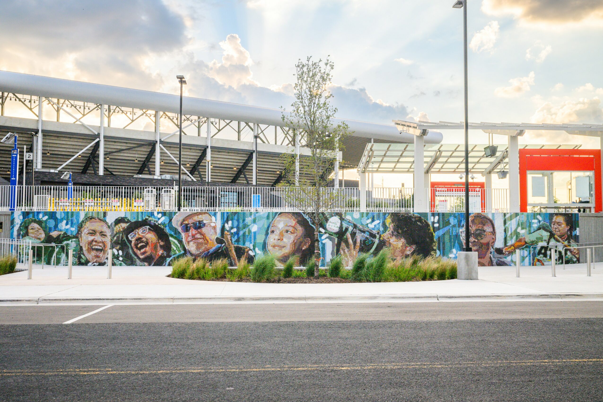 Colorful mural of diverse people's faces on a wall, with a stadium and sky in the background.