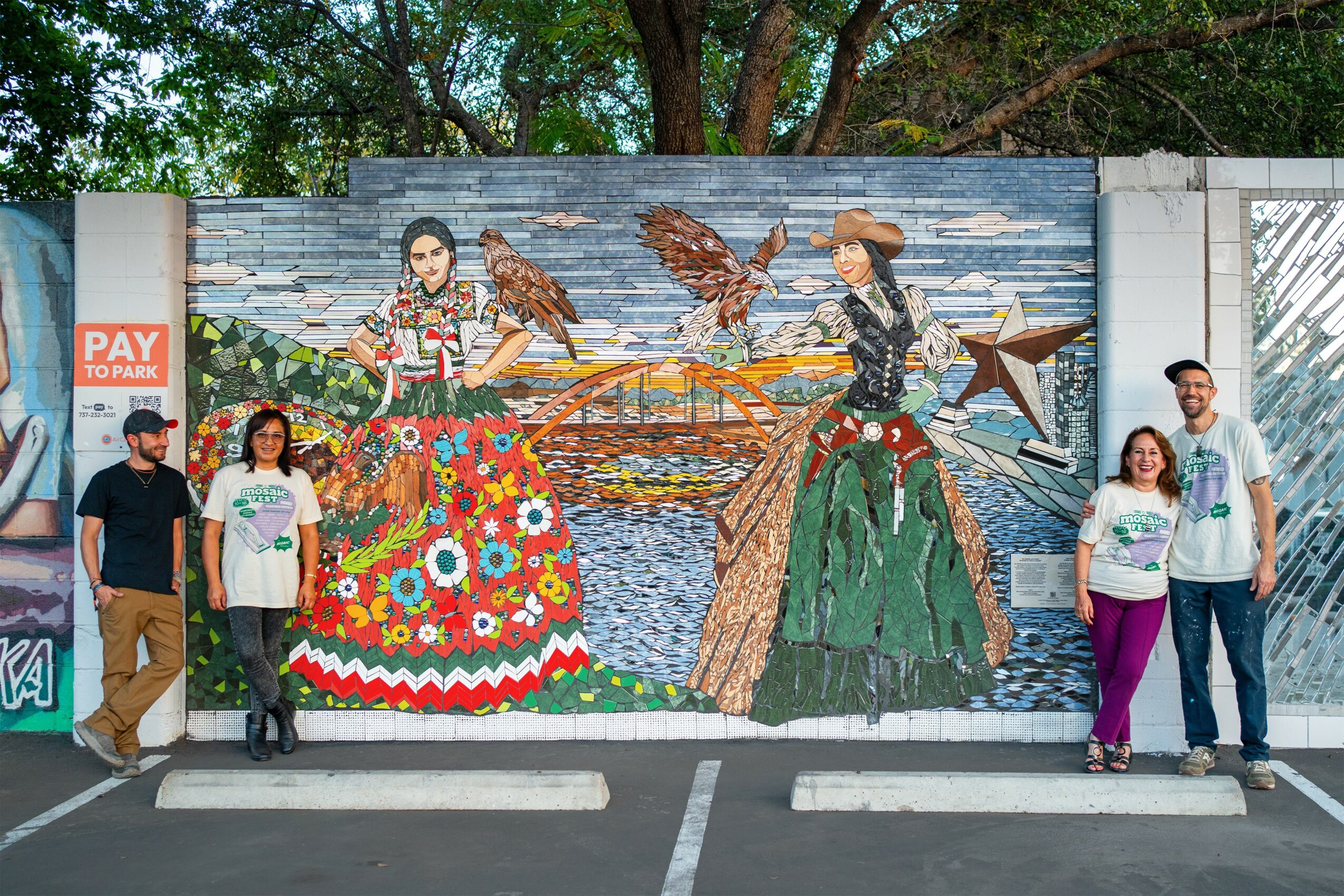 Four people stand in front of a colorful mural depicting two women in traditional dresses with birds, trees, and water background.