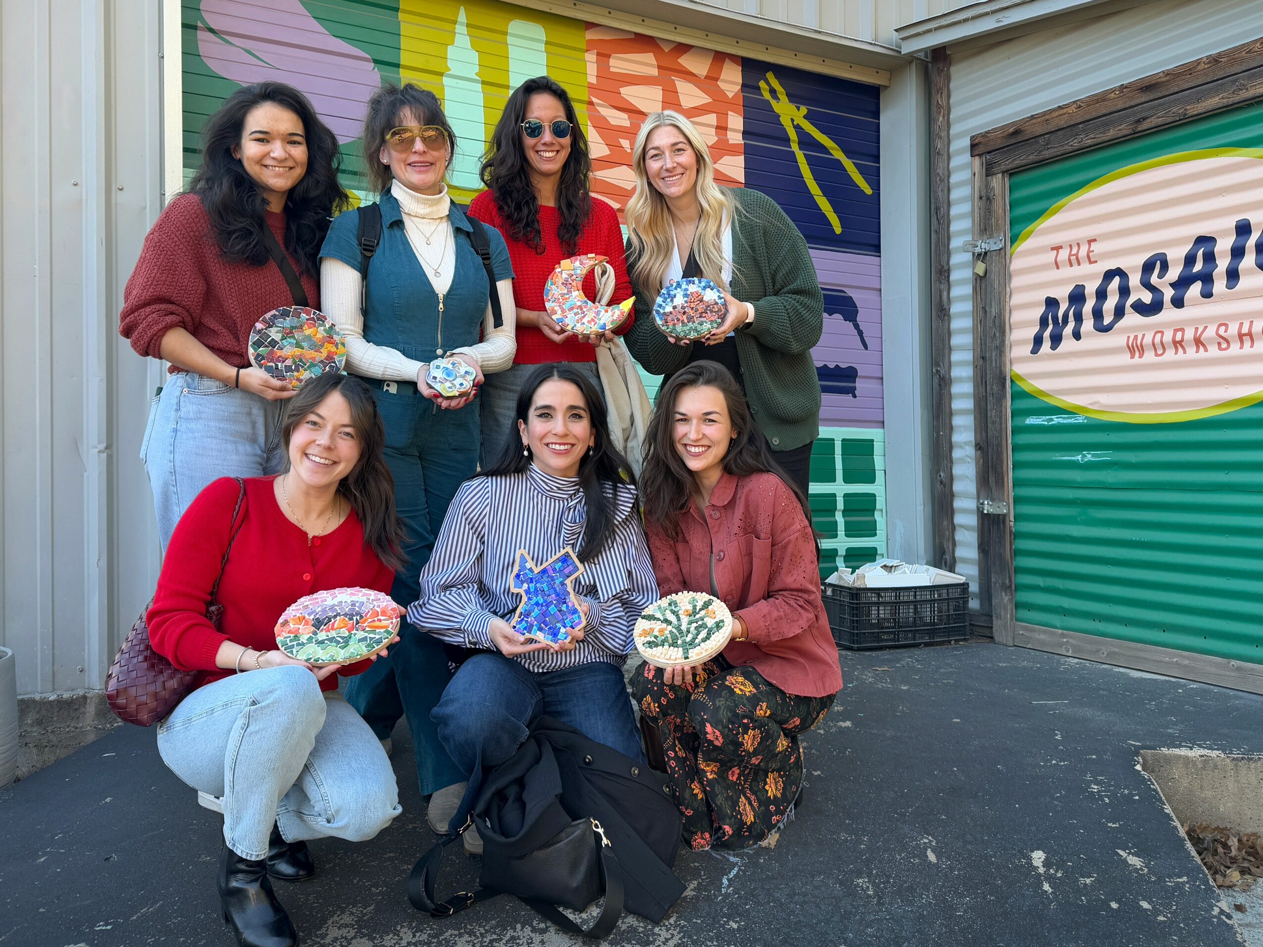 Seven people holding decorated objects stand in front of colorful mural and a closed shop with a sign. Group smiling.
