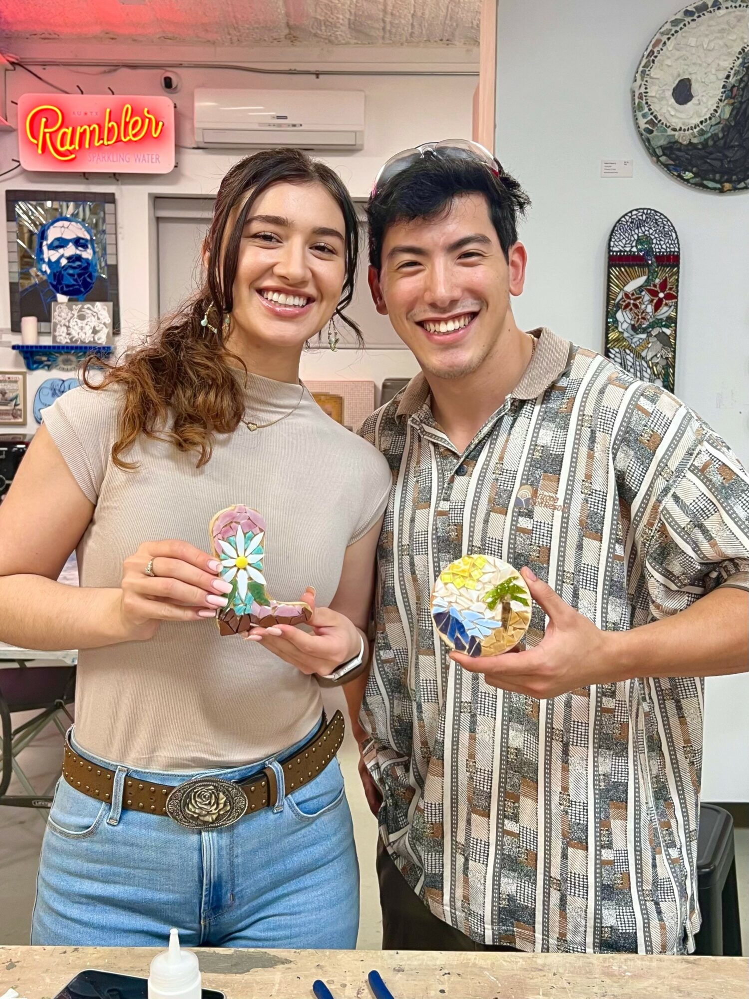 Two smiling people holding small decorative objects indoors, with a neon sign and wall art in the background.