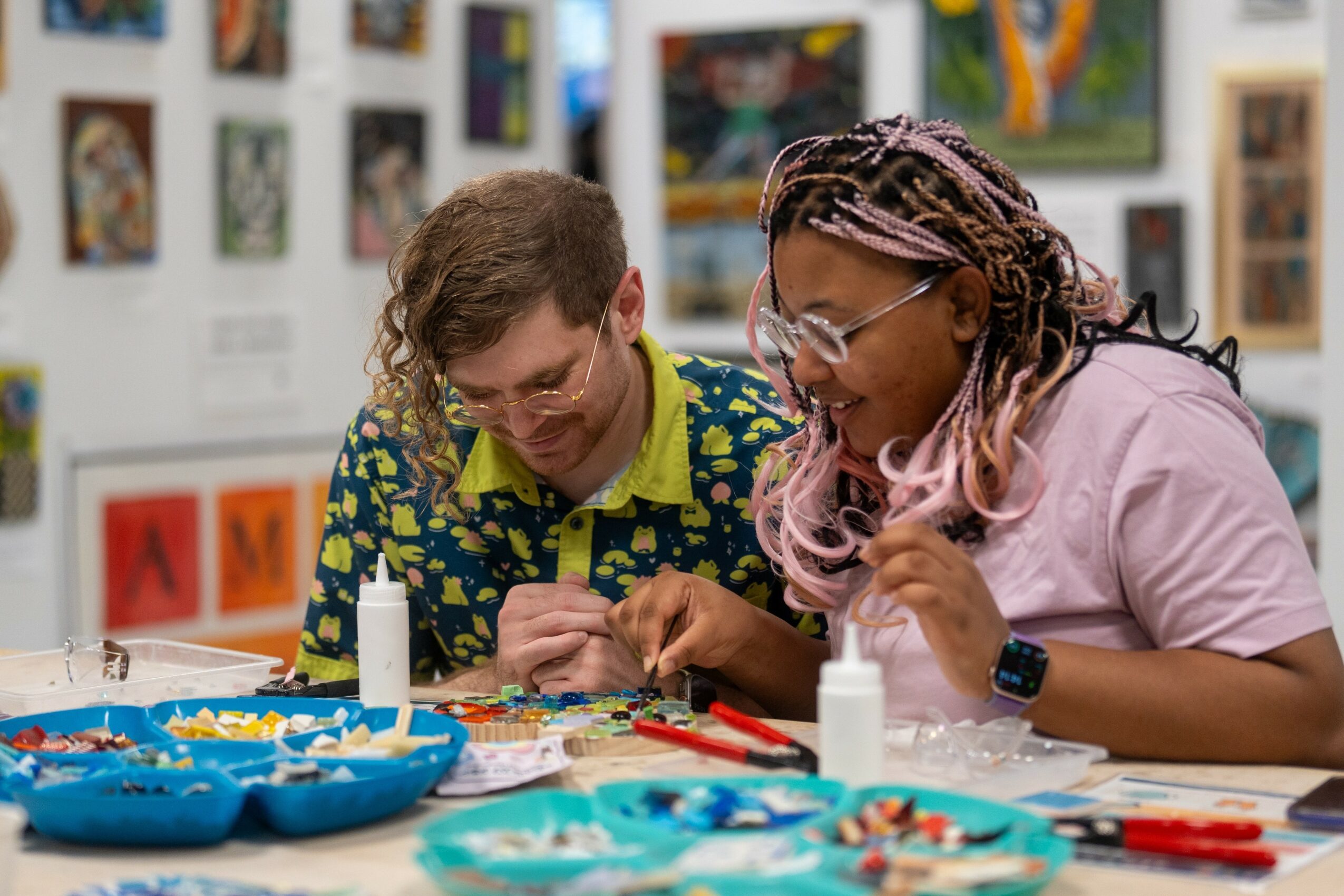 Two women working on an art project at a table with art supplies, in an art gallery setting.