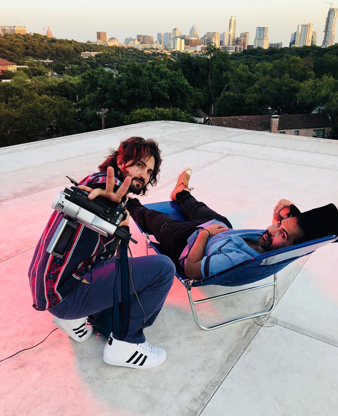 Two people relaxing on a rooftop with a city skyline in the background, one sitting and the other lying on a lounge chair.