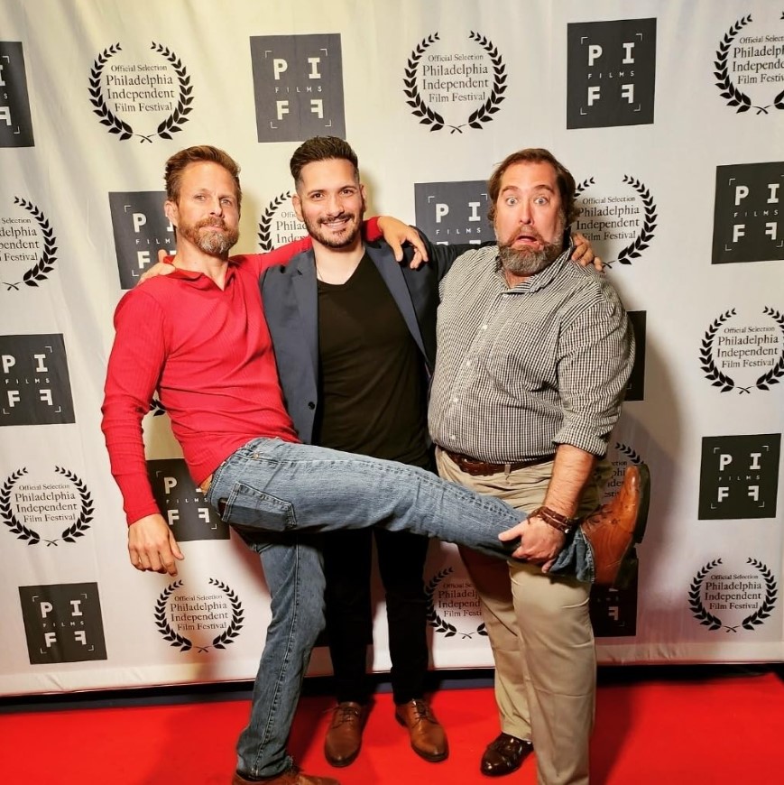 Three men posing on a red carpet in front of a backdrop with logos and laurels, one holding a shoe, smiling.