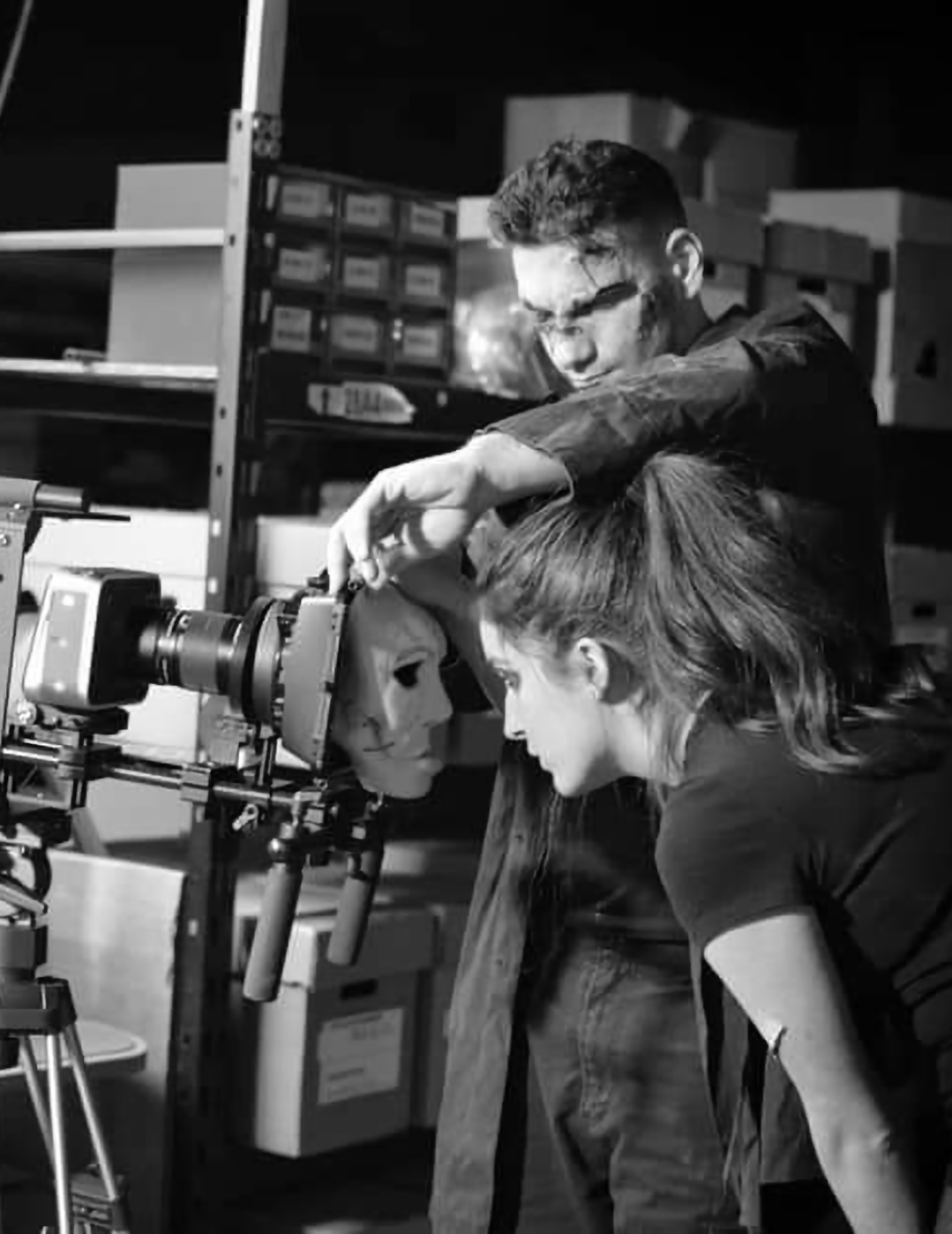 Two people working with a camera or filming equipment in a storage room or workshop, shelves with boxes in background.
