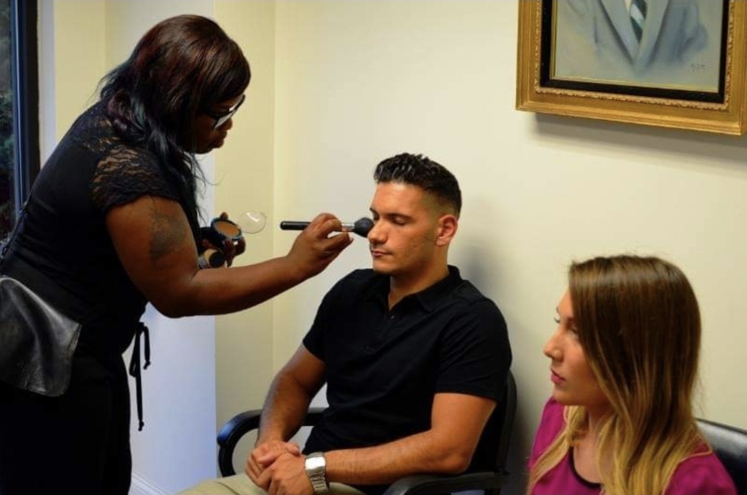 Makeup artist applying makeup to a seated man, with a woman sitting nearby in a room with a framed picture on the wall.