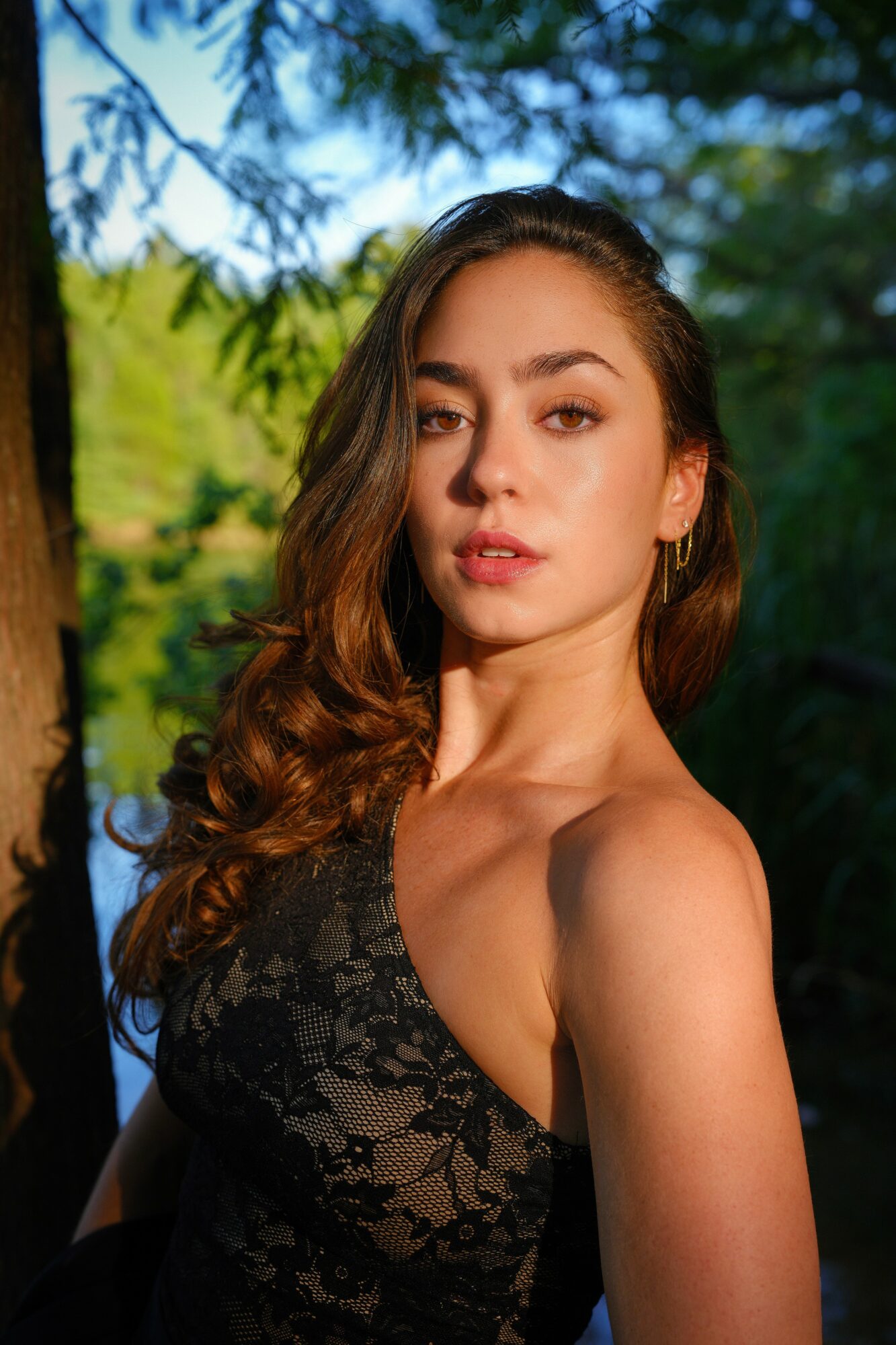 Young woman with long, wavy hair outdoors, wearing a black lace top, surrounded by green trees and blue sky.