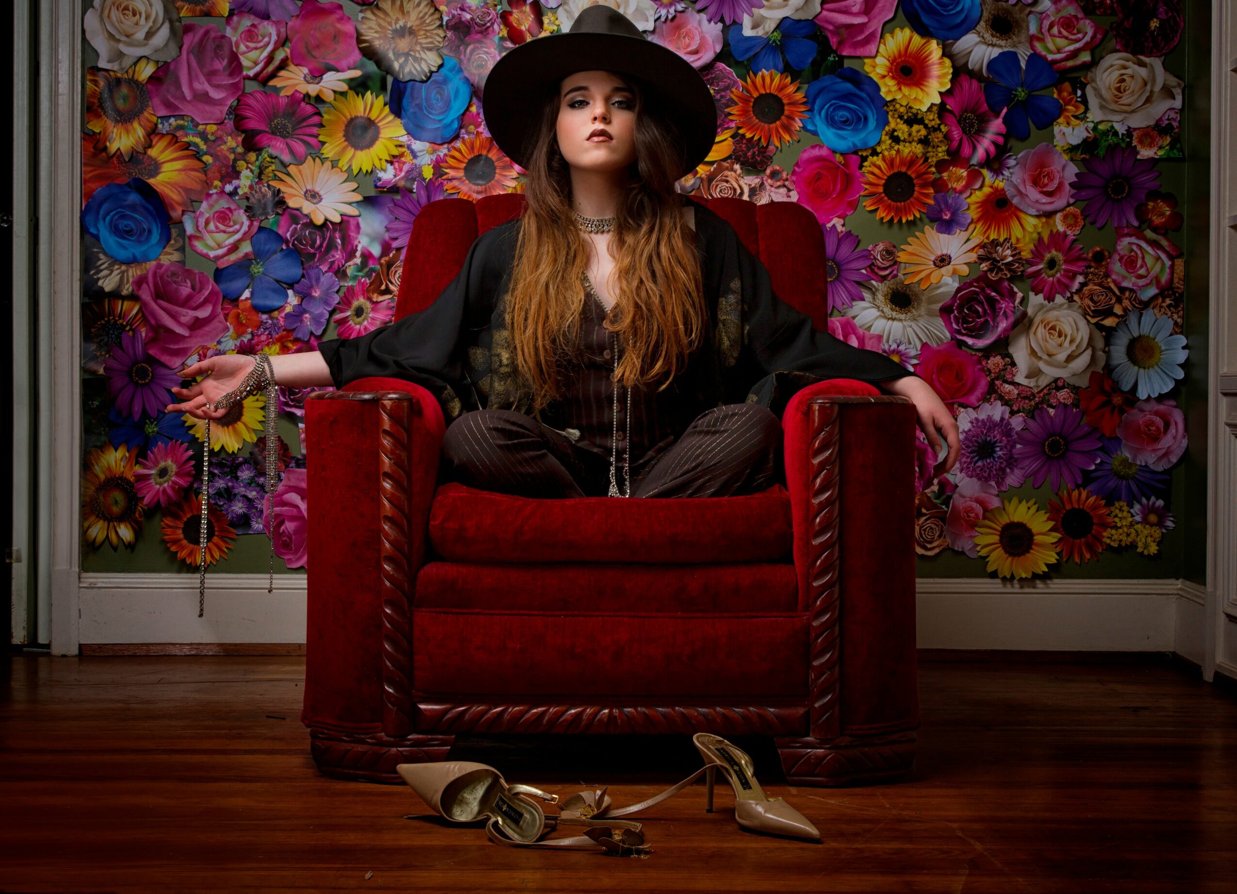 Woman sitting on a red armchair with a floral wall background, wearing a black hat and outfit, with shoes and a belt on the floor.
