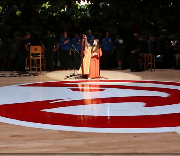 Two women in traditional dresses stand on a stage with microphones, surrounded by people, with a large red and white symbol on the floor.