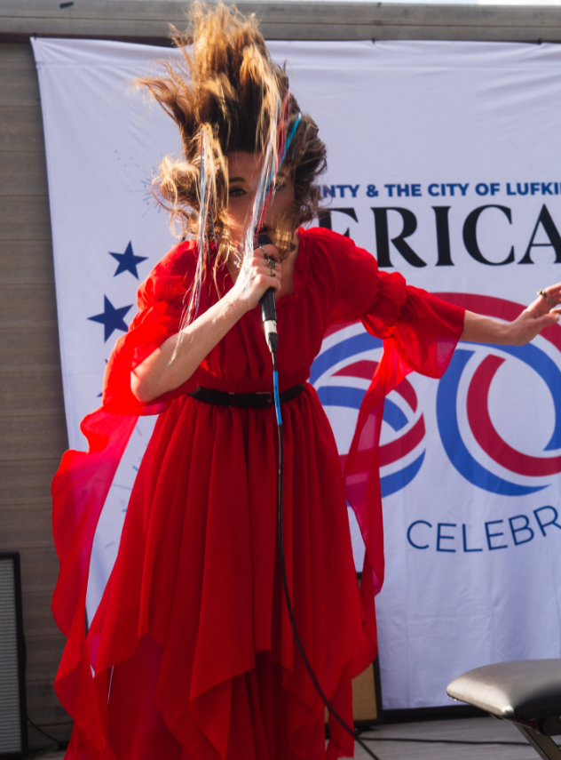 Person with long hair wearing a red dress, holding a microphone, hair flying, standing in front of a banner.