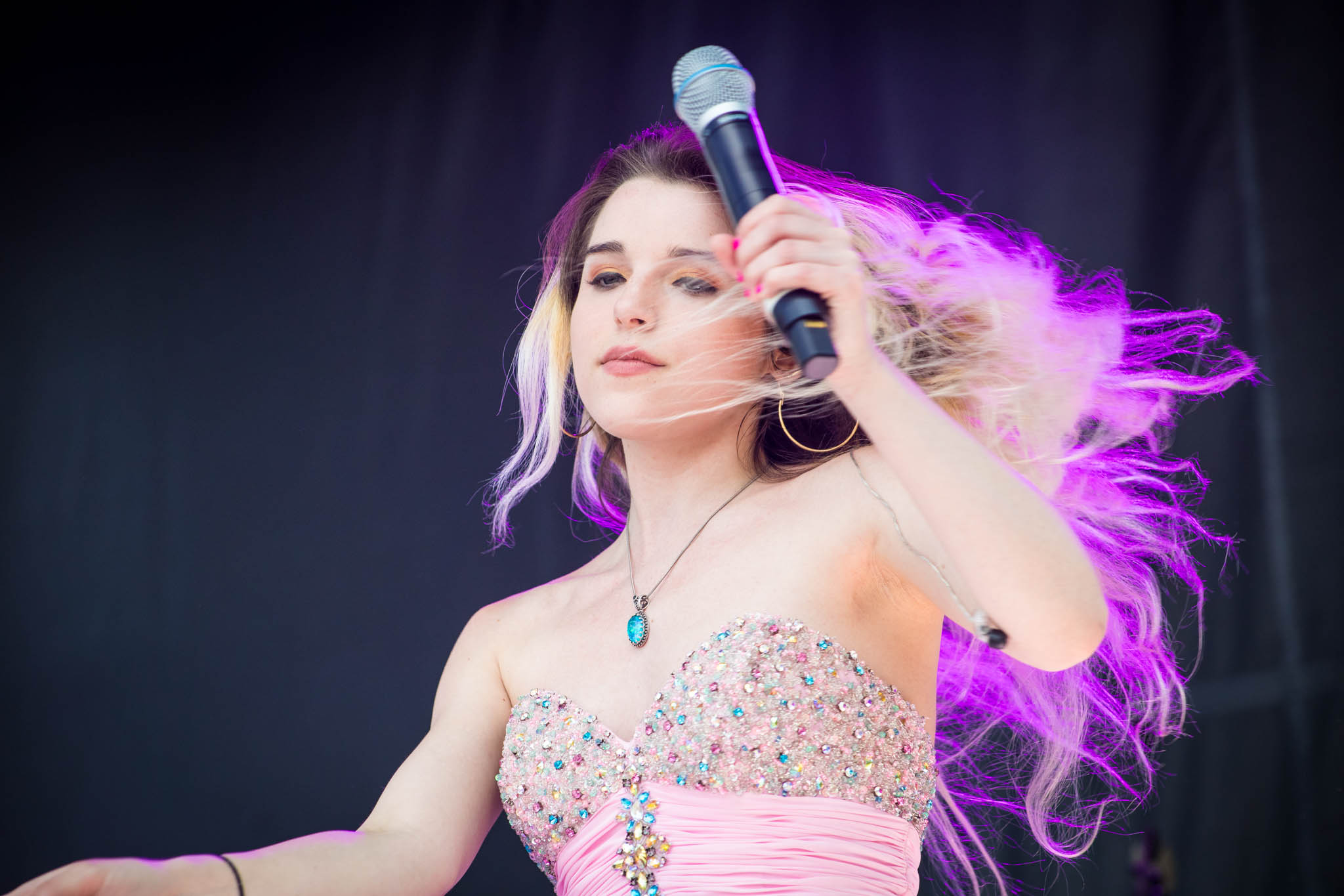 Young woman singing into a microphone on stage with pink lighting in her hair, wearing a sparkly strapless top and necklace.