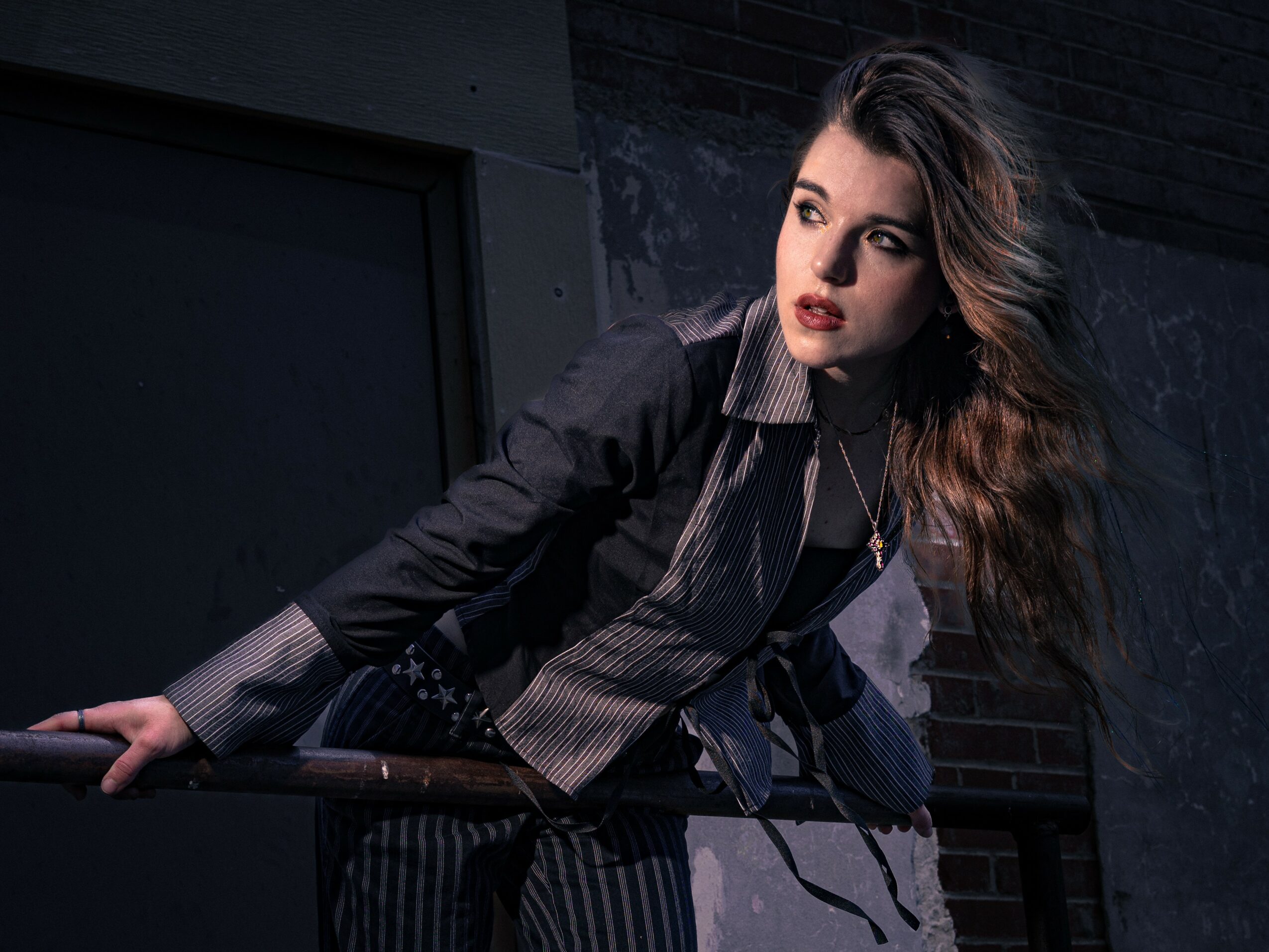 Woman with long hair leaning on a railing, wearing dark clothing and a scarf, in an urban setting.