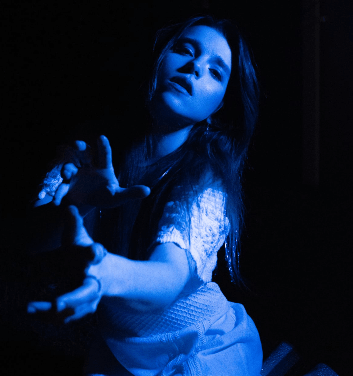 Young woman with long hair reaching out, illuminated by blue light, in a dark setting.
