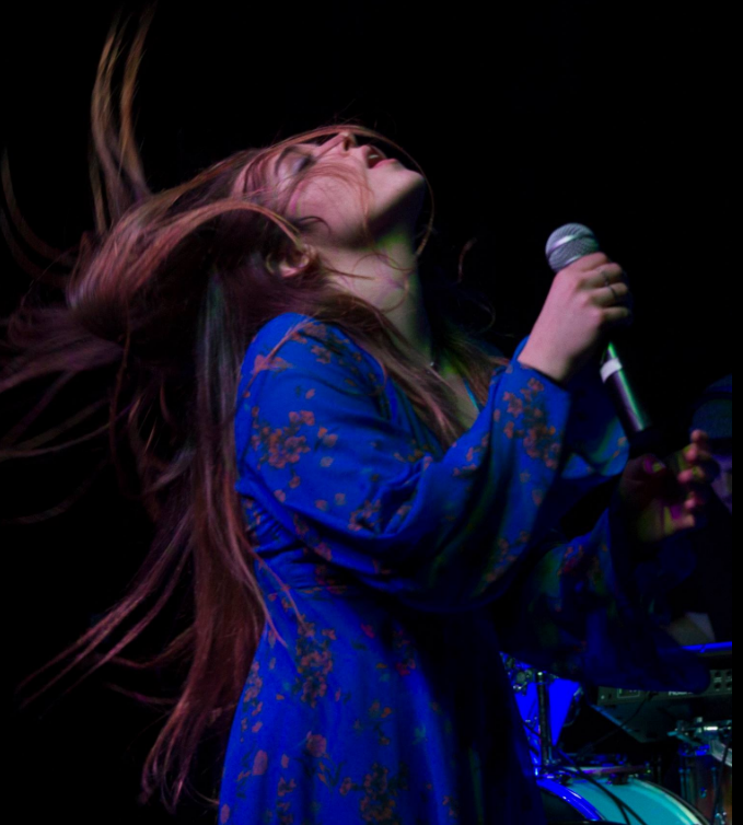 Person singing into a microphone with long hair, wearing a blue patterned garment, on a dark stage.